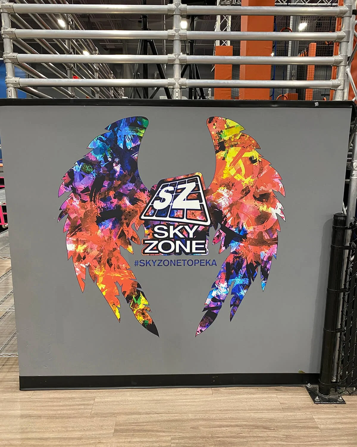 Colorful eagle wings graphic with the logo 'SZ SKY ZONE' and the hashtag #SKYZONEtoPEKA on a gray background at Sky Zone trampoline park.