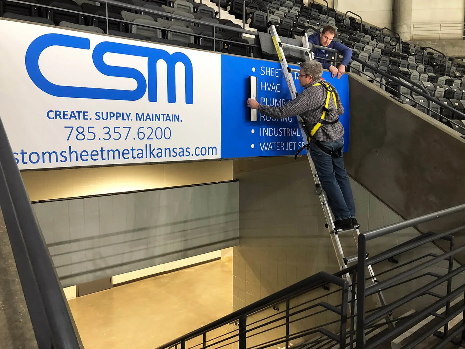 Two men installing a large blue sign on an arena wall, one on a ladder and another on a platform. The sign displays the logo and contact information for a metal sheet company, with services listed including HVAC, plumbing, roofing, industrial, and wa