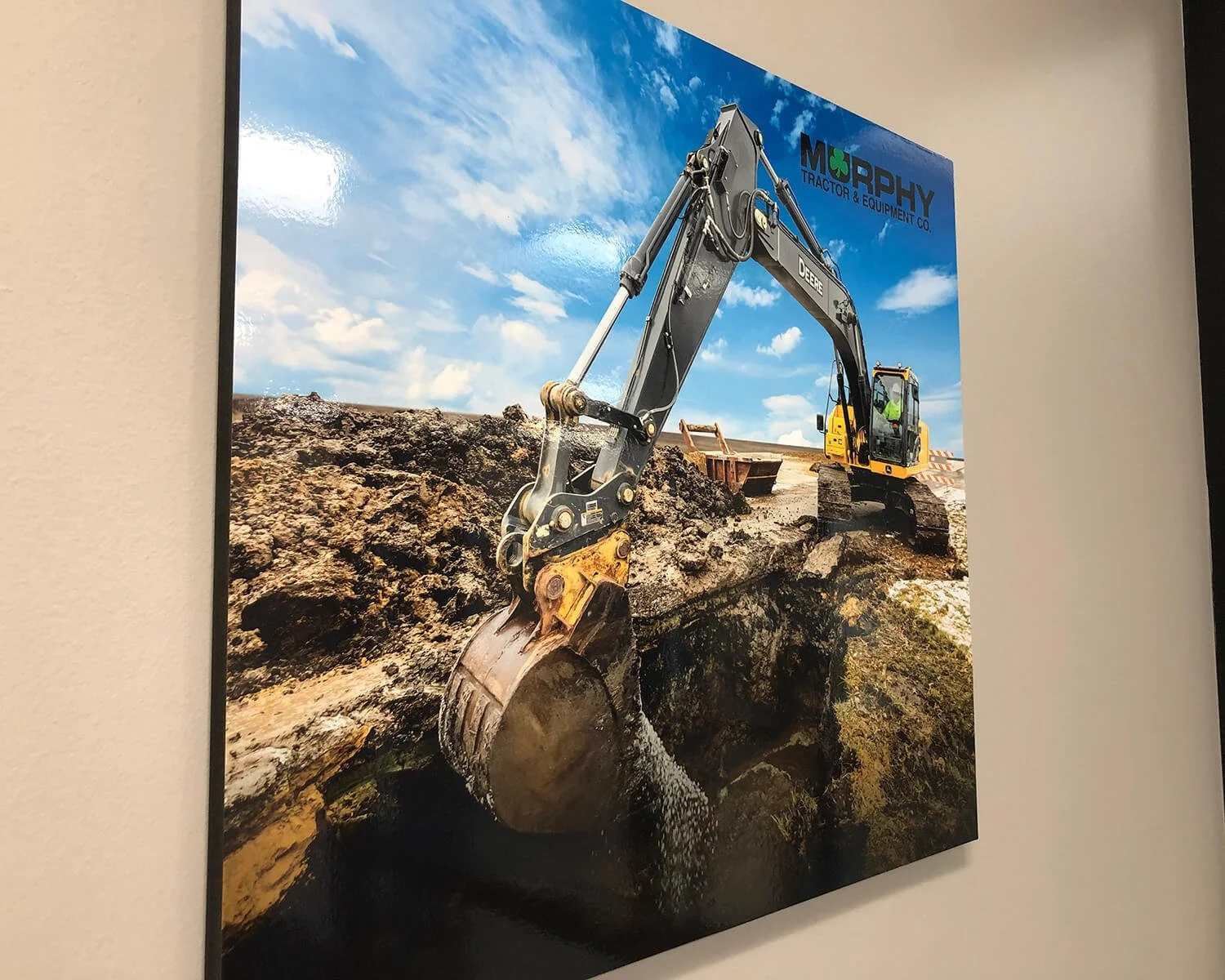 Photograph of an excavator digging into the ground on a construction site, with blue sky and clouds in the background. The brand 'DEERE' is visible on the excavator arm, and a sign with 'MORPHY TRACTOR & EQUIPMENT CO.' is in the upper right corner of