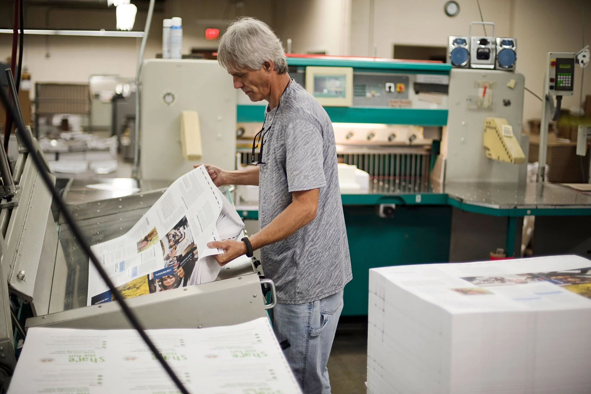 A man in casual clothing operating printing machinery in a print shop, inspecting printed materials.
