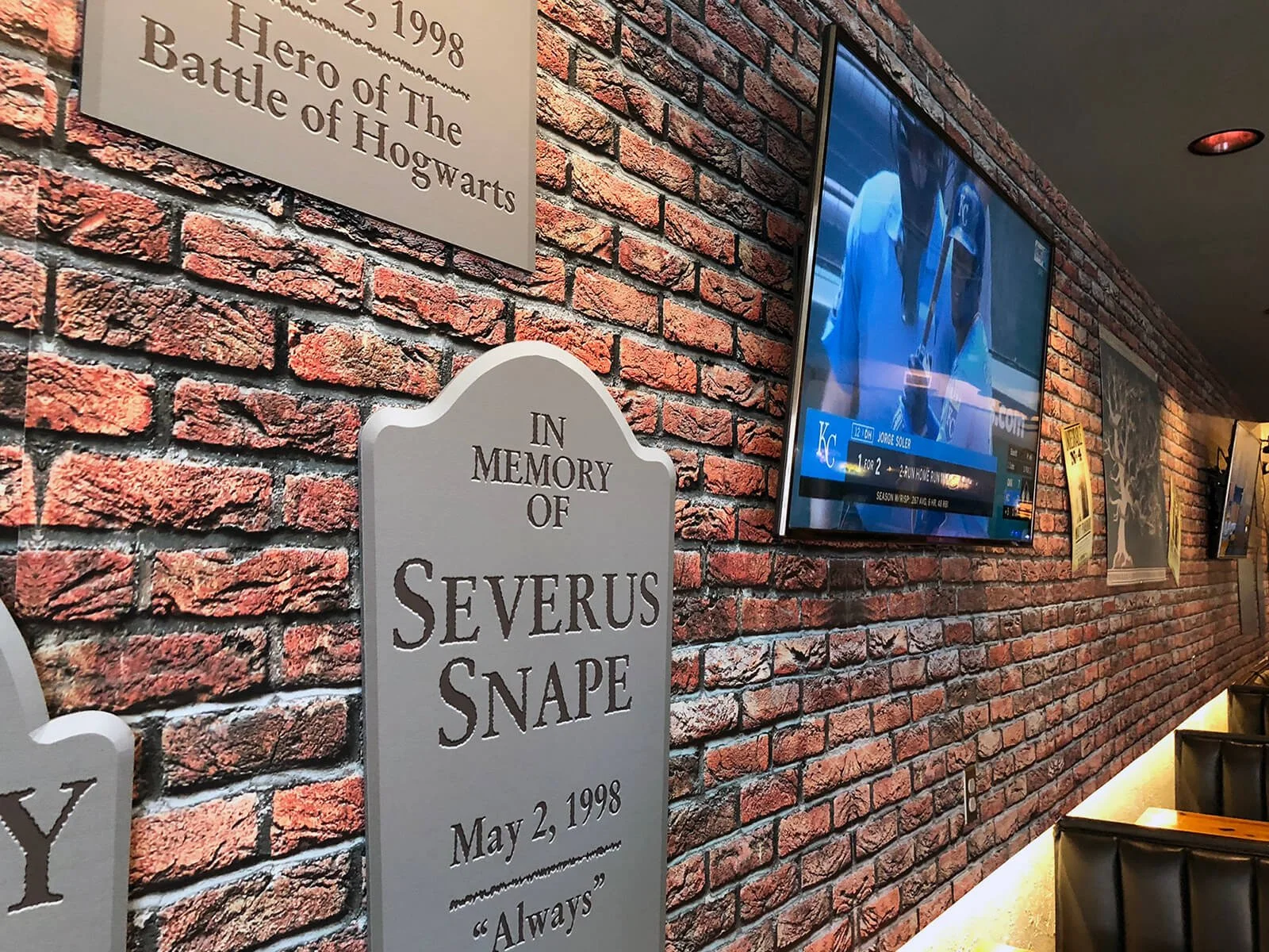 A brick wall decorated with memorial plaques and a mounted television. One plaque reads: 'In Memory of Severus Snape, May 2, 1998, Always.' A television screen displays a sports broadcast.
