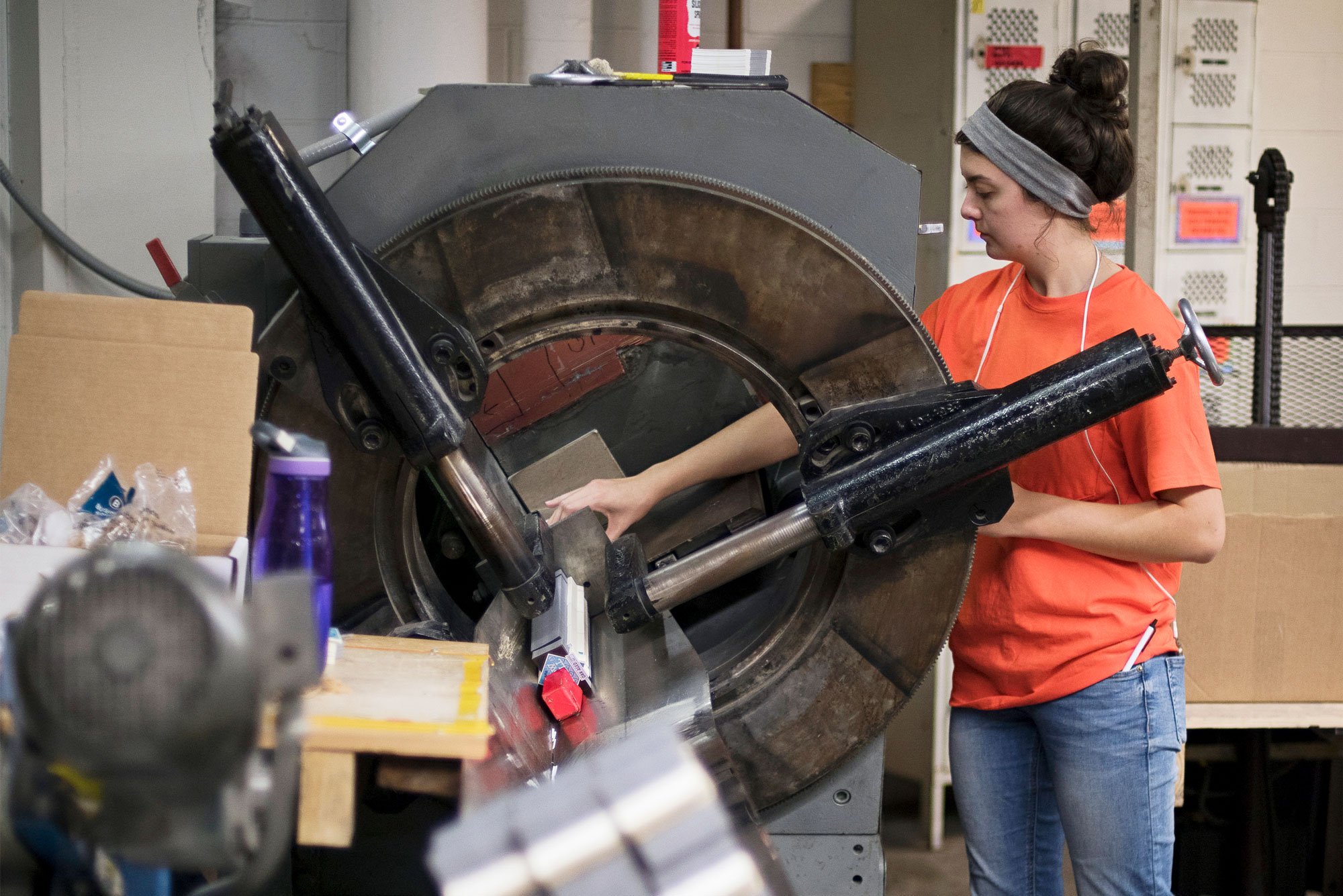 A woman working with a large industrial machine in a factory or workshop setting, wearing an orange shirt and a gray headband.