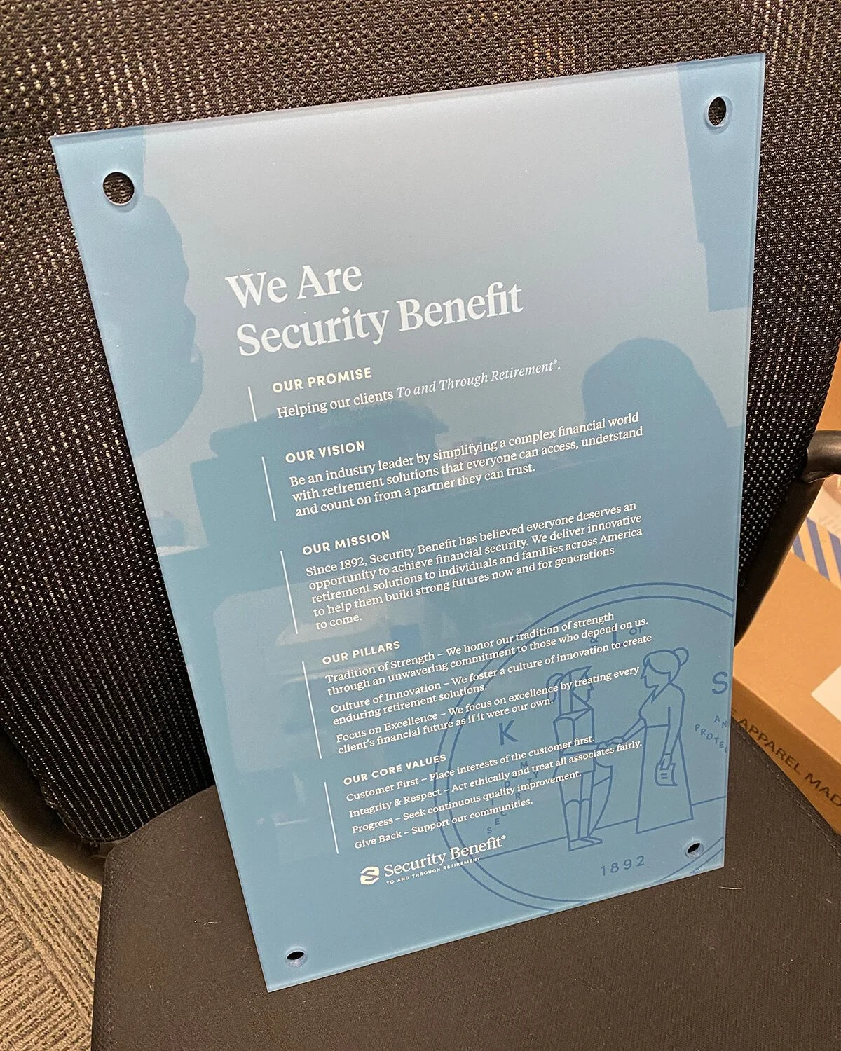 Light blue printed poster with white text and a line drawing of two women at a vending machine, leaning on it and holding shopping bags, on a black cushioned chair. The poster describes Security Benefit's mission, vision, promise, pillars, and core v