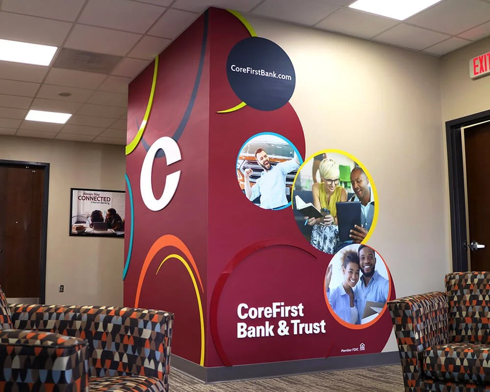 Corner of a bank lobby with a colorful wall display. The display features images of diverse people working or collaborating and the text 'CoreFirst Bank & Trust' and 'CoreFirstBank.com'. There are patterned chairs in the foreground and a framed poste