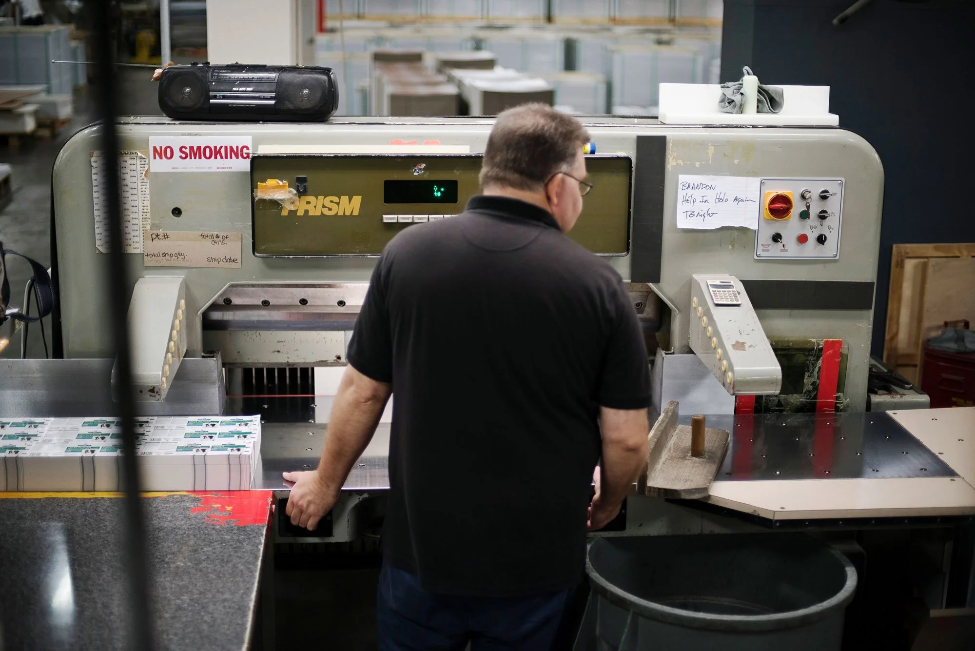 A man in a black shirt working in a factory or warehouse, standing in front of a large industrial machine.