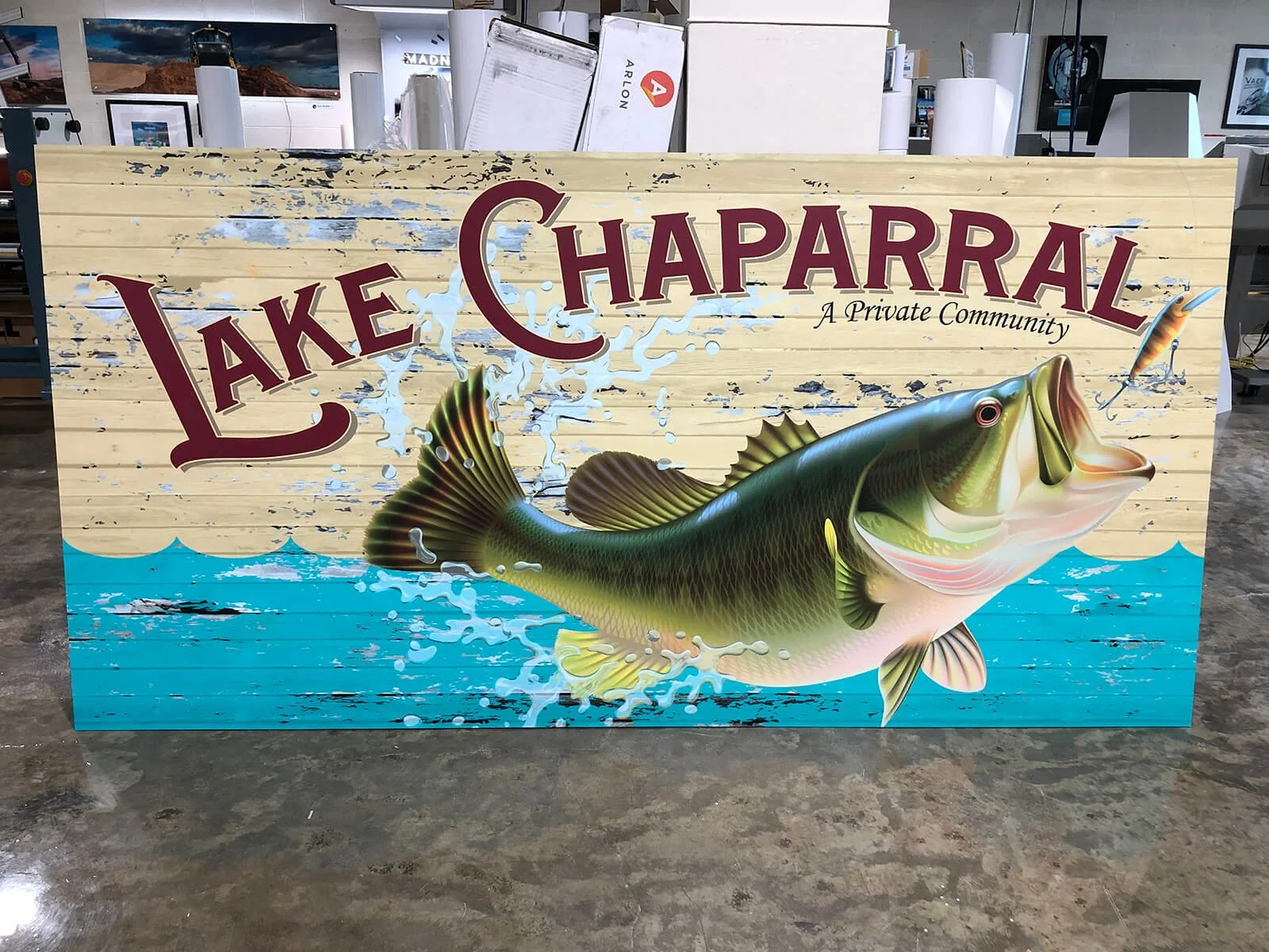 A colorful sign for Lake Chaparral, a private community, showing a large bass fish jumping out of blue water, with the community name in large red letters and a background that resembles weathered wood.
