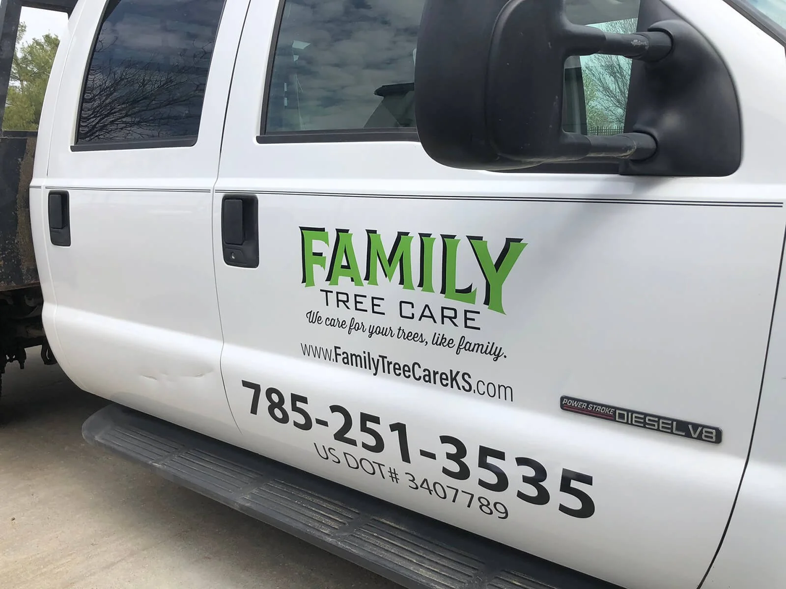 White truck with 'Family Tree Care' logo and contact information on the door, parked on a concrete surface, with trees and sky reflected on the window.