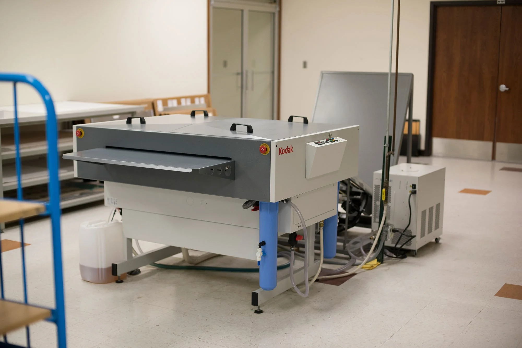 A large laboratory machine with the brand Kodak, connected to various tubes and devices, situated in a room with chairs and closed doors.