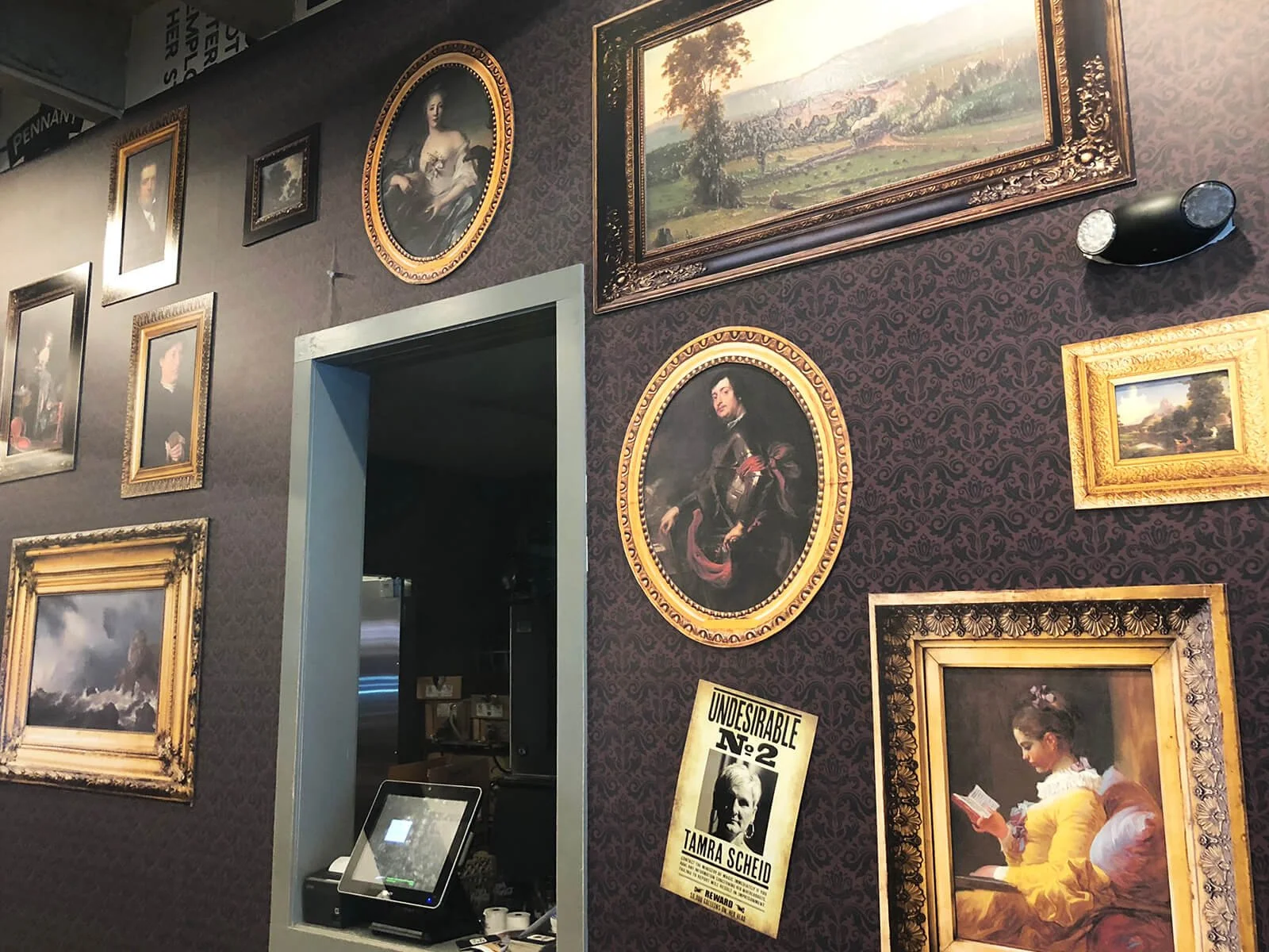 Wall decorated with various framed paintings, portraits, and landscapes, with a door showing a glimpse into an interior space and a promotional poster for Tamra Scheid.
