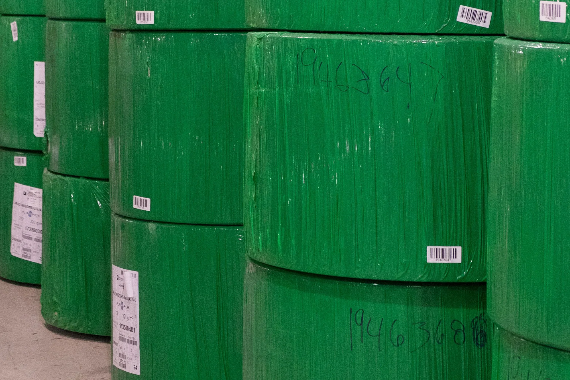 Large green cylindrical rolls stacked in a storage area, with barcode labels and handwritten numbers on their surface.