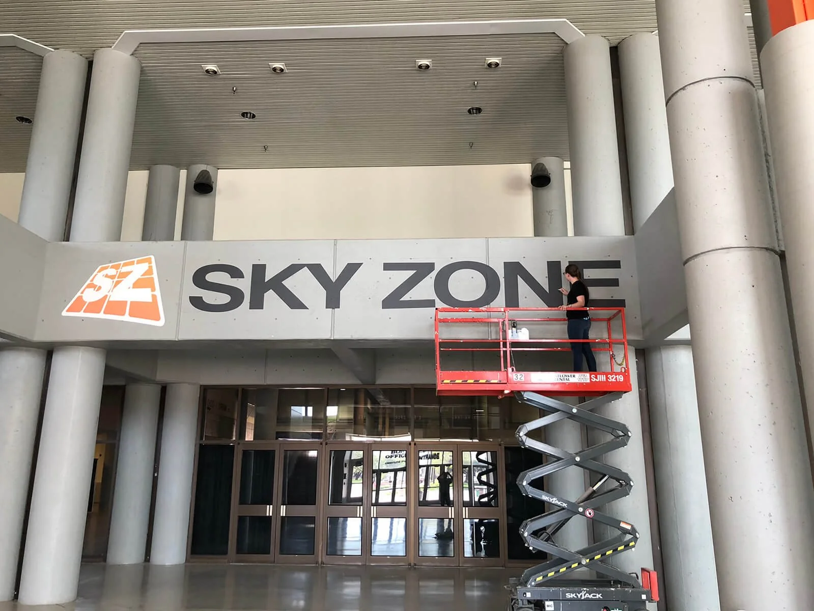 A person on a scissor lift installing a large sign that reads 'SKY ZONE' at an indoor trampoline park.