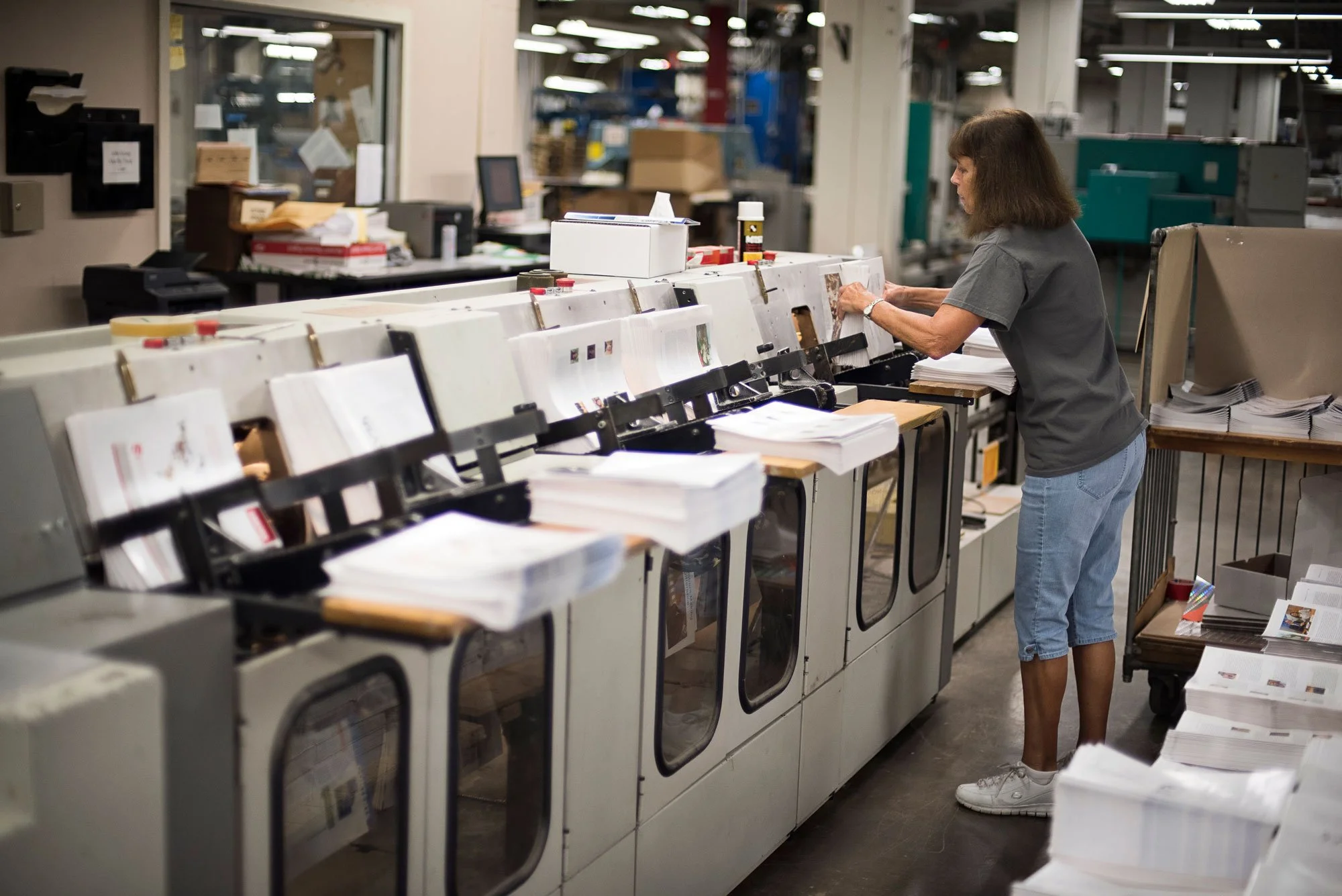 A woman working on a printing or binding machine in a busy industrial or print shop setting, surrounded by stacks of paper and published materials.