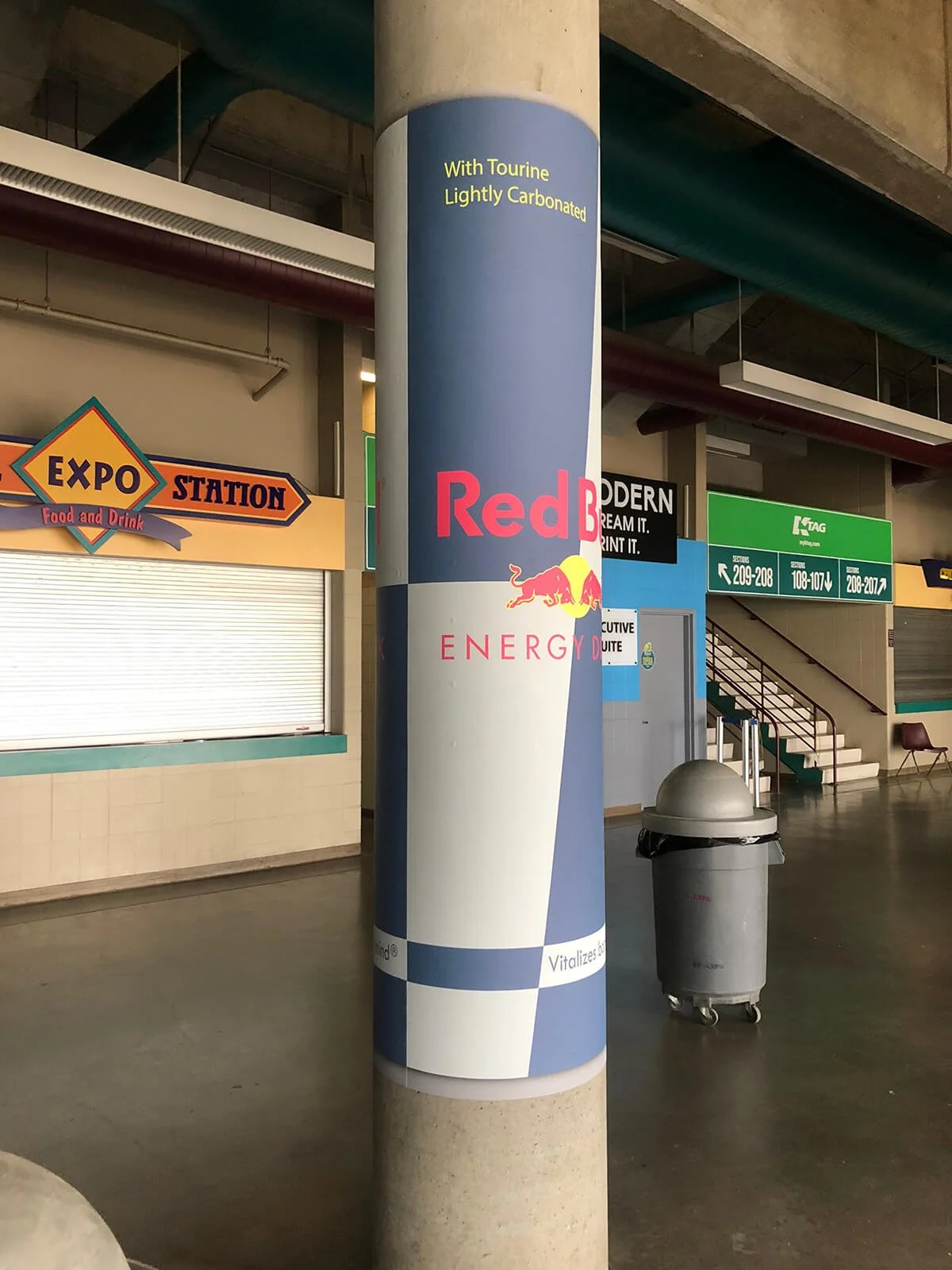 A cylindrical advertisement sign for Red Bull Energy Drink with the text 'With Tourine Lightly Carbonated'. In the background, there is a food and drink stand with a sign that reads 'EXPO STATION', and a trash bin.