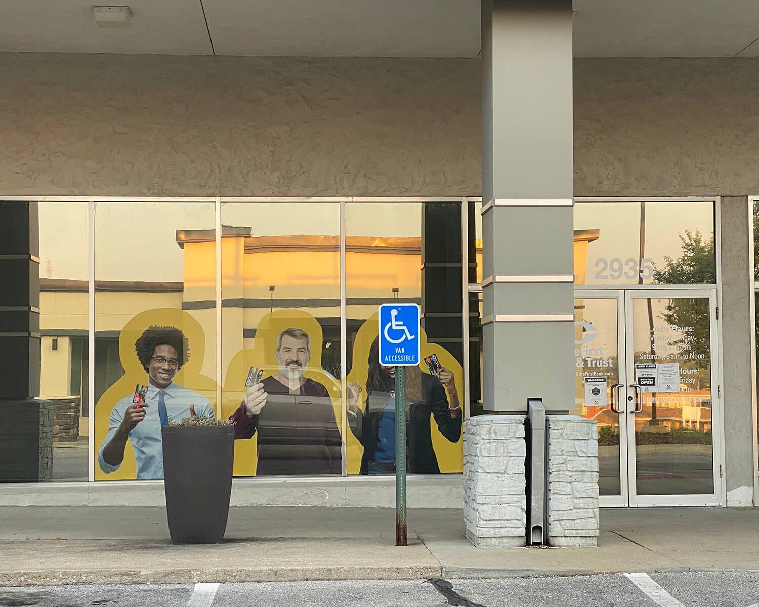 The storefront features a large window with life-size cutouts of diverse smiling people holding credit cards, with a blue accessible parking sign in front. The building number is 2935, and the door has signage indicating business hours.