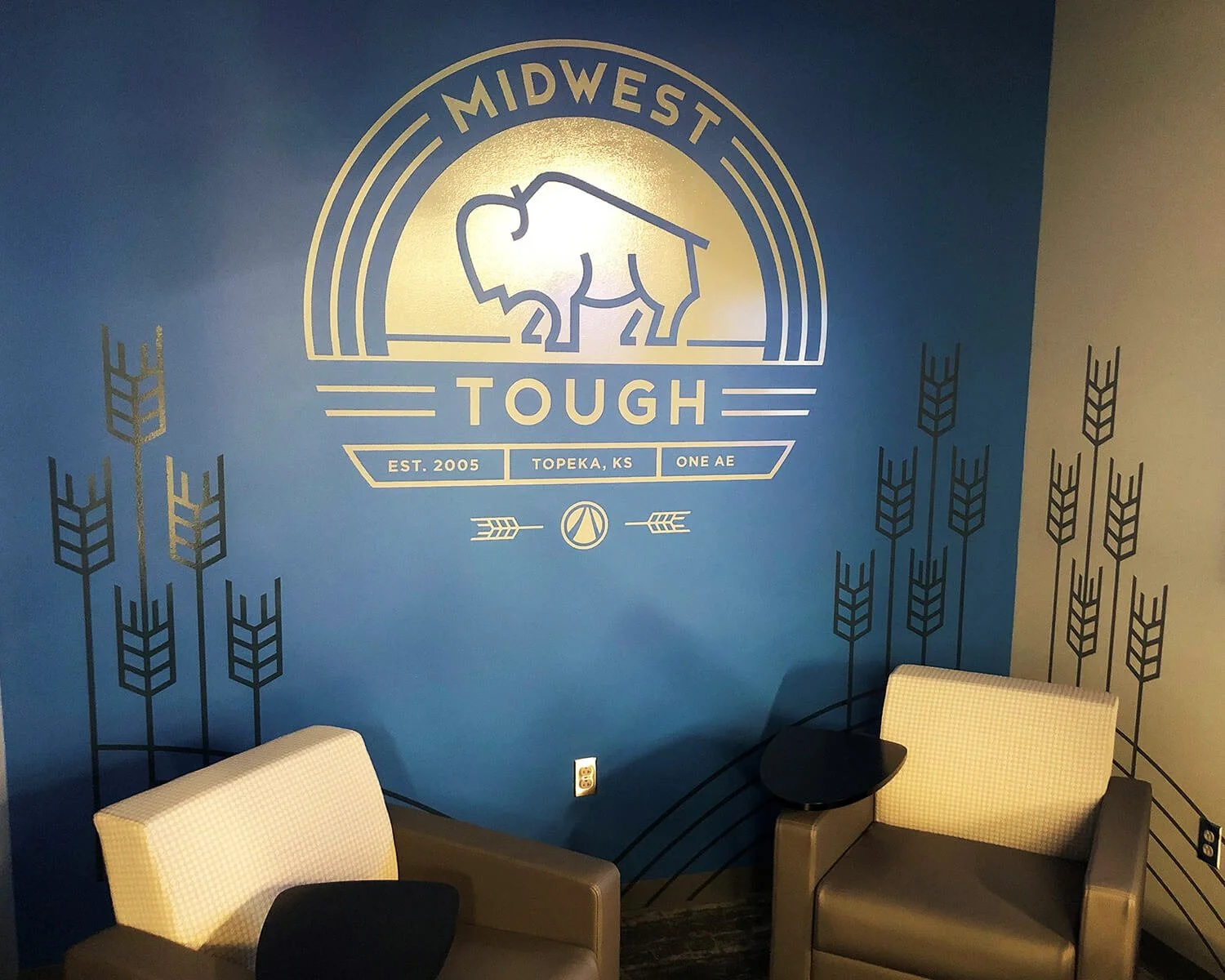 Interior of a room with a blue wall featuring a logo of a buffalo and the words 'Midwest Tough.' Two beige armchairs are positioned against the wall with a small black side table between them.