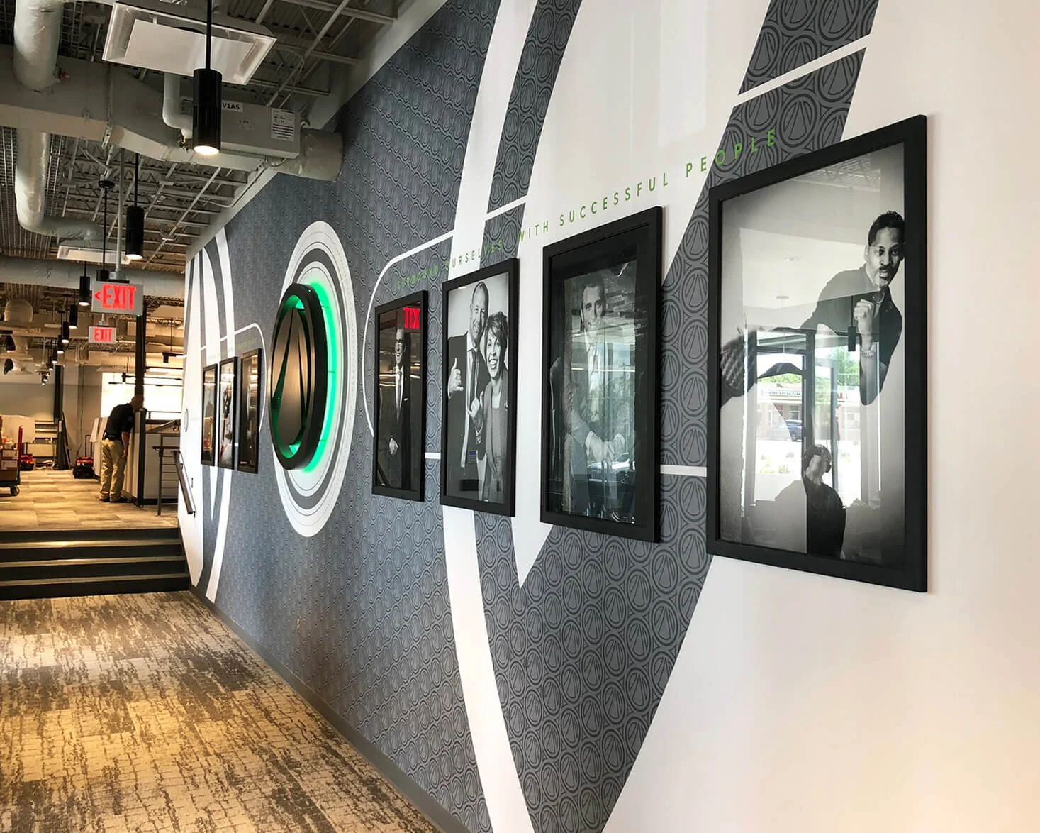 Office hallway with framed photographs of diverse people and a logo on the wall. Green text reads 'Yourself with successful people.'