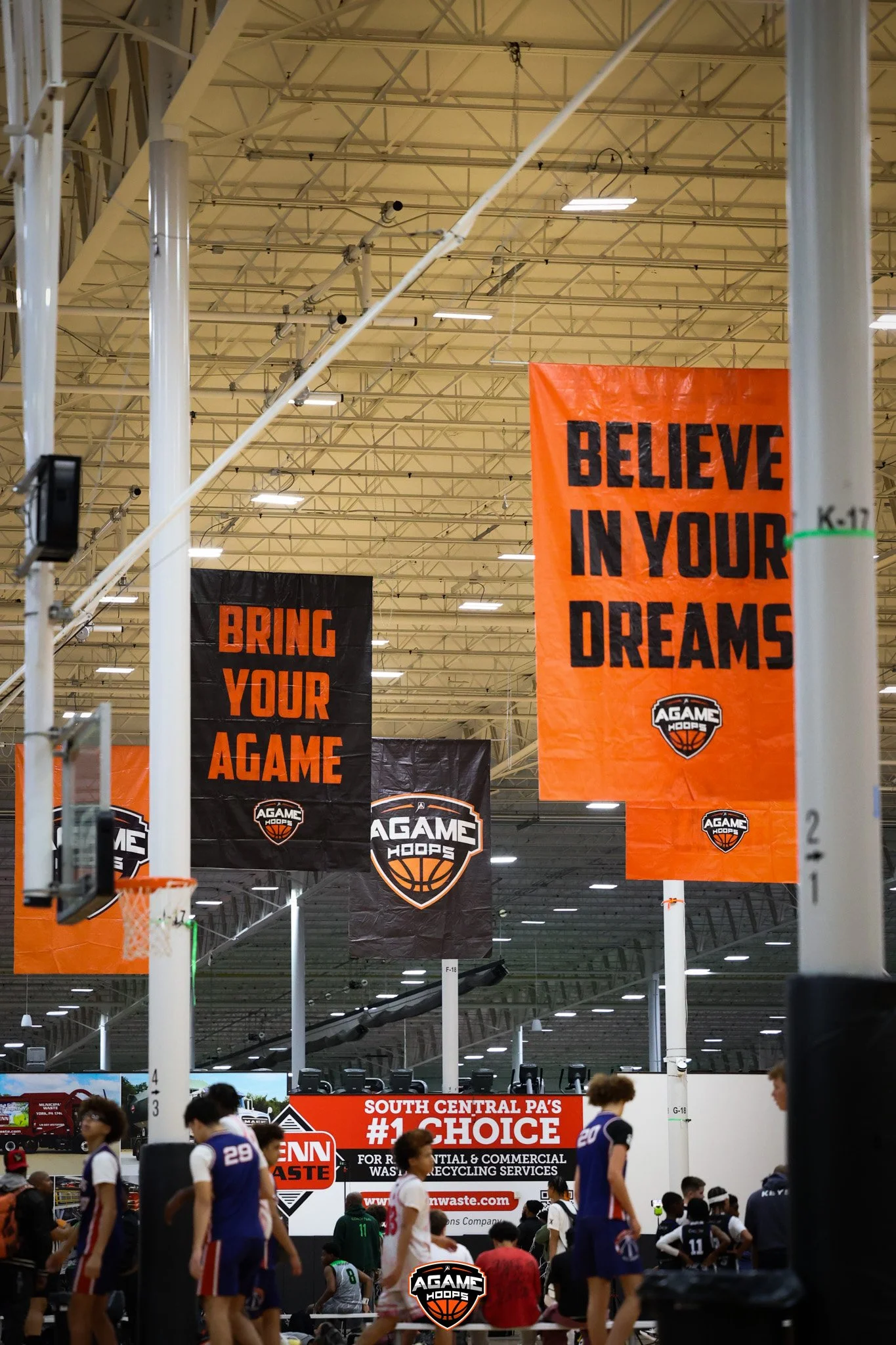 Indoor basketball court with players and banners hanging from the ceiling, including an orange banner with the text 'BELIEVE IN YOUR DREAMS' and black banners with the text 'BRING YOUR A GAME'.