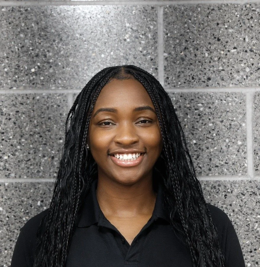 A young woman with braided hair smiling in front of a gray, speckled cinder block wall.