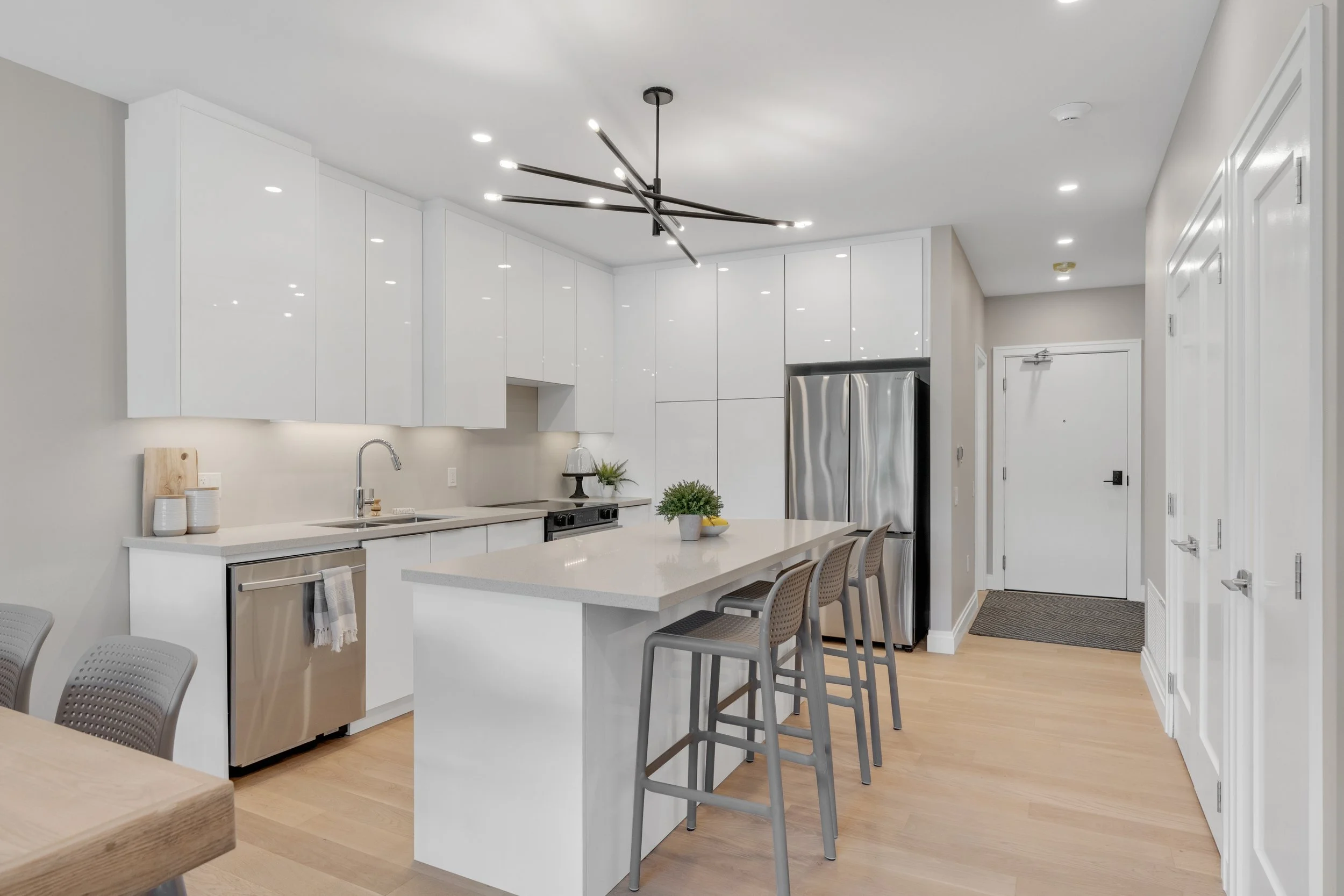 Modern kitchen with white cabinets, a large island, a stainless steel refrigerator, and bar stools, illuminated by ceiling lights and a contemporary black chandelier.  The Riverbend Condos, Huntsville, Muskoka.