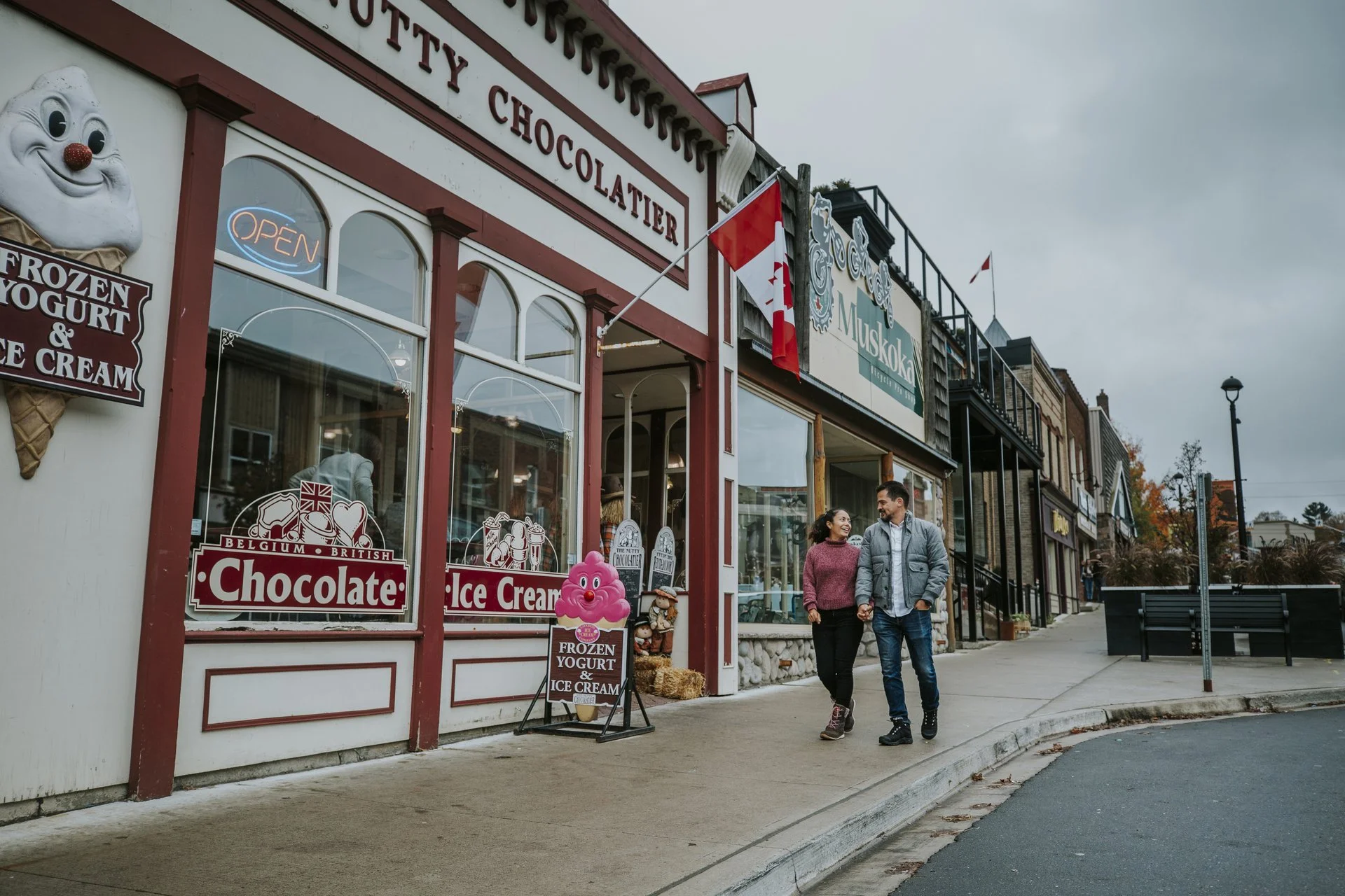A street scene with two people walking and smiling outside a chocolate shop. The shop has signs for ice cream, frozen yogurt, and Belgium and British chocolates.  The Riverbend Condos, Huntsville, Muskoka.