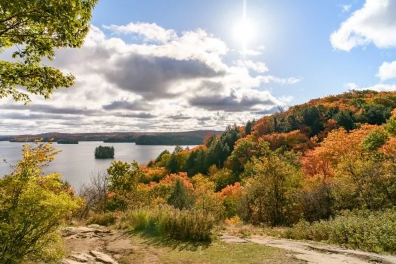 Scenic view of a lake surrounded by colorful autumn foliage and a partly cloudy sky with sunlight breaking through. The Riverbend Condos, Huntsville, Muskoka.
