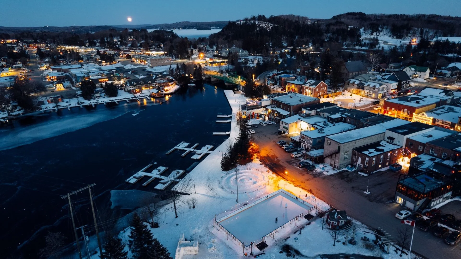 An aerial view of a snowy small town at dusk with Christmas lights, a frozen lake with docks, and a skating rink surrounded by snow.  The Riverbend Condos, Huntsville, Muskoka.