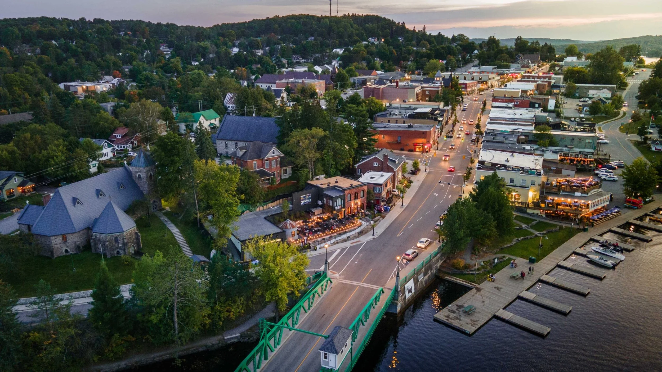 Aerial view of a small waterfront town at sunset with a bridge, marina, historic church, and bustling main street lined with shops and restaurants.  The Riverbend Condos, Huntsville, Muskoka.