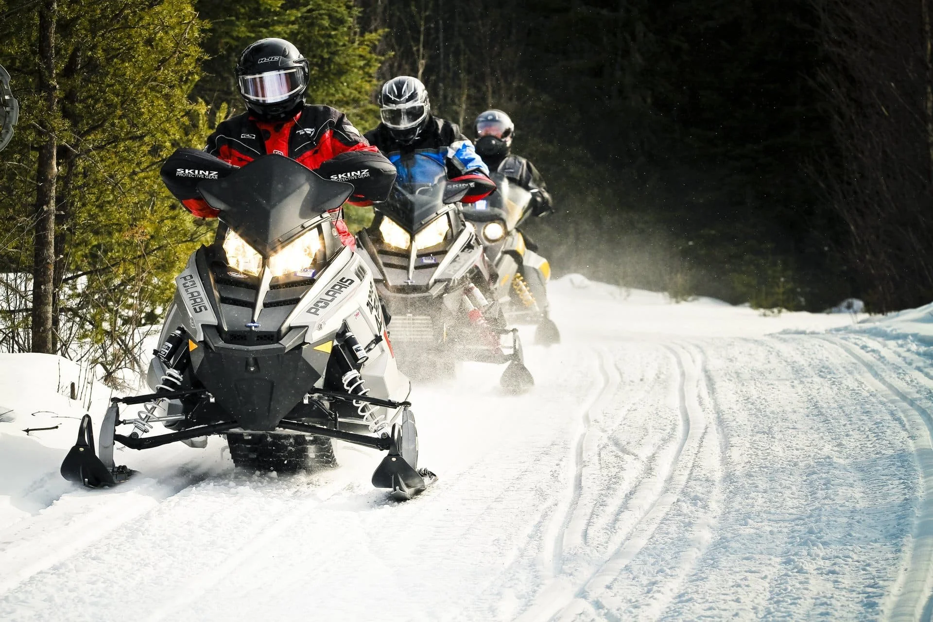 Group of four people riding snowmobiles on a snow-covered trail through a forest.  The Riverbend Condos, Huntsville, Muskoka.