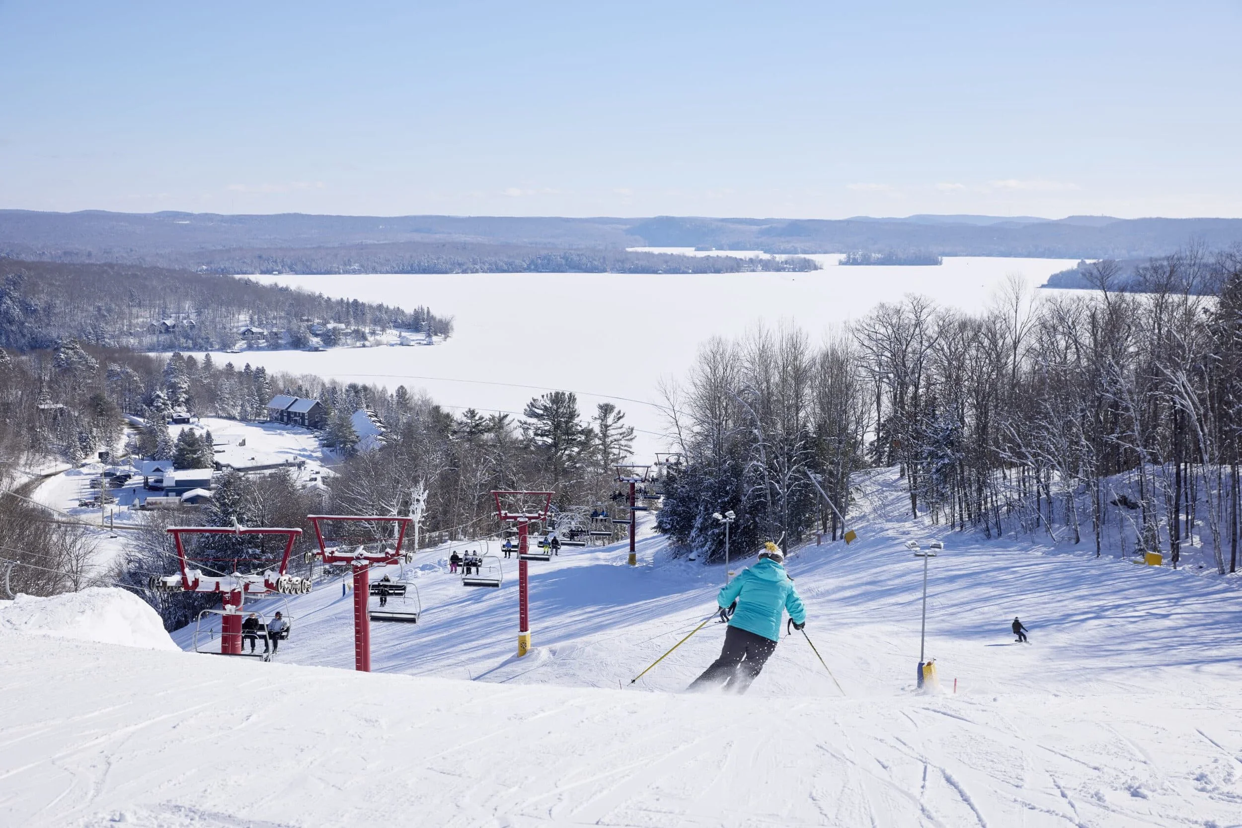 A person skiing downhill on a snow-covered slope with ski lifts, leafless trees, and a frozen lake in the background on a clear winter day.  The Riverbend Condos, Huntsville, Muskoka.