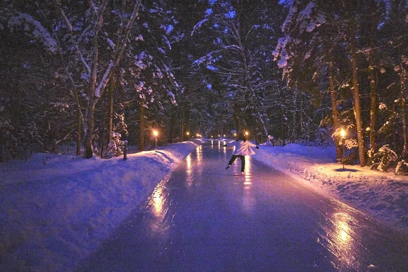 People ice skating on a snow-covered path lined with snow-laden trees at night, illuminated by small street lamps.  The Riverbend Condos, Huntsville, Muskoka.