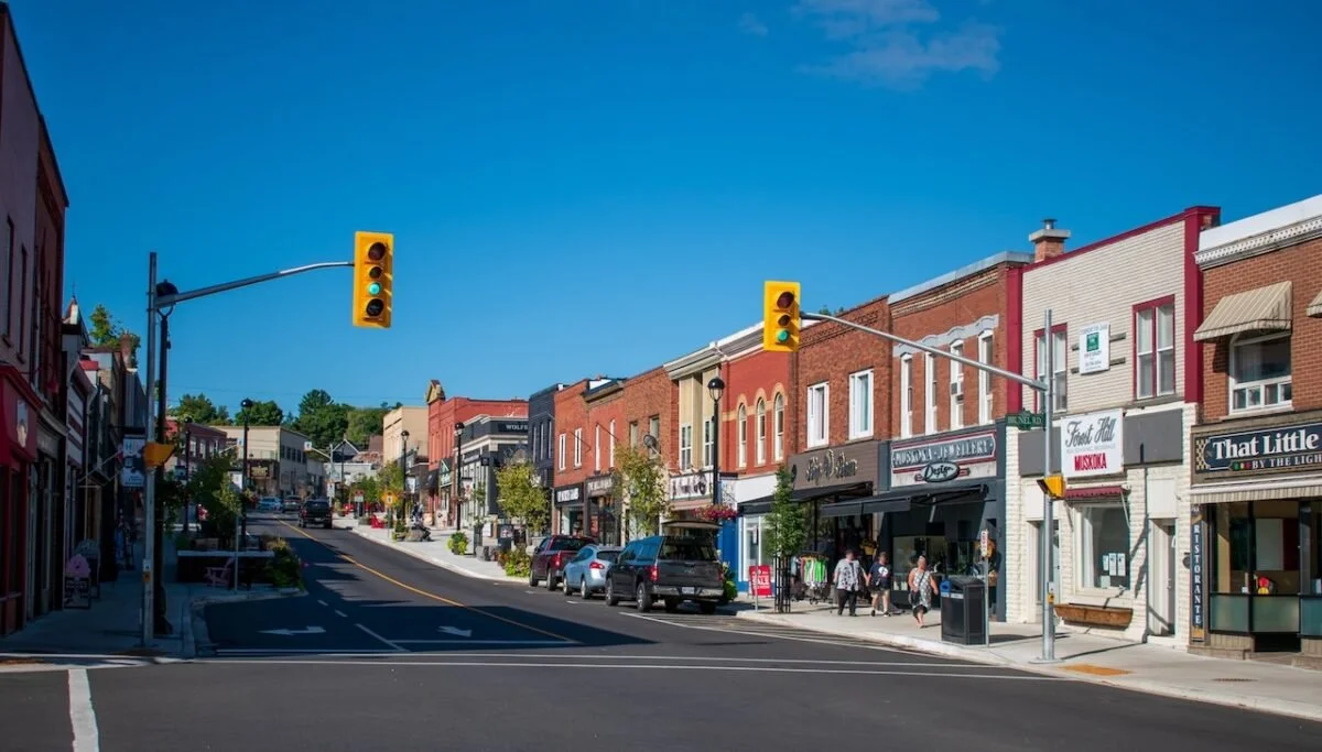 A small downtown street with parked cars, shops, and pedestrians under a clear blue sky.  The Riverbend Condos, Huntsville, Muskoka.