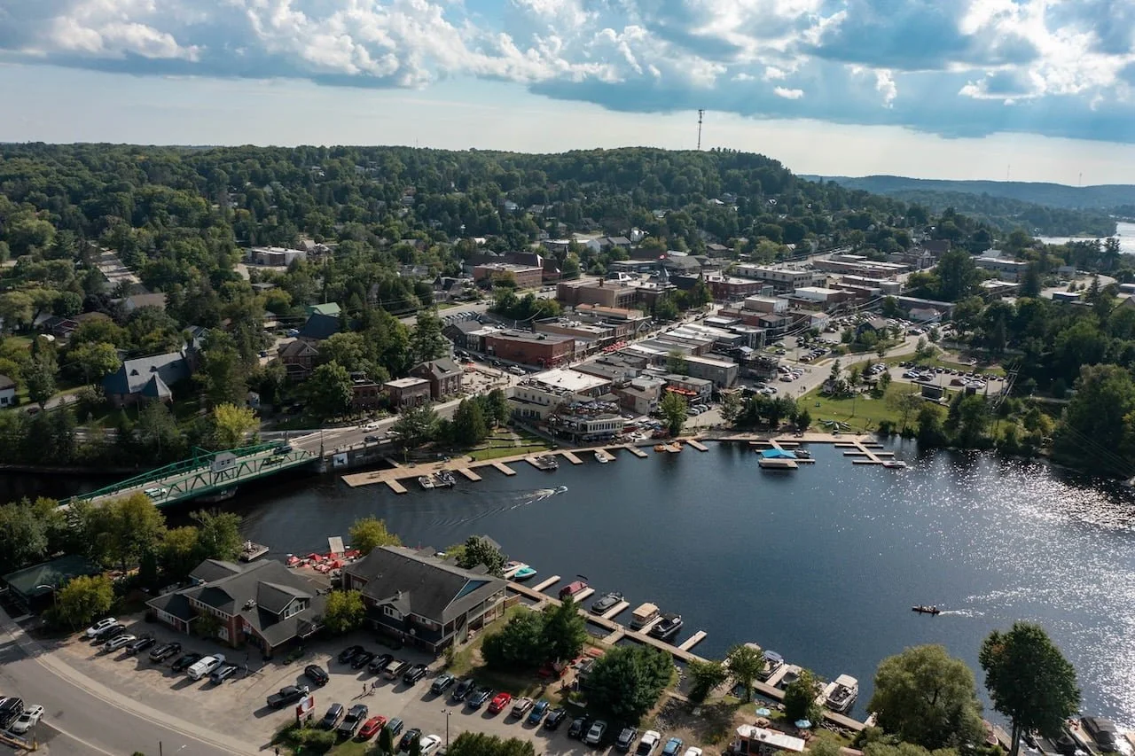 Aerial view of a small town near a body of water with boats and docks, surrounded by trees and hilly landscape.  The Riverbend Condos, Huntsville, Muskoka.