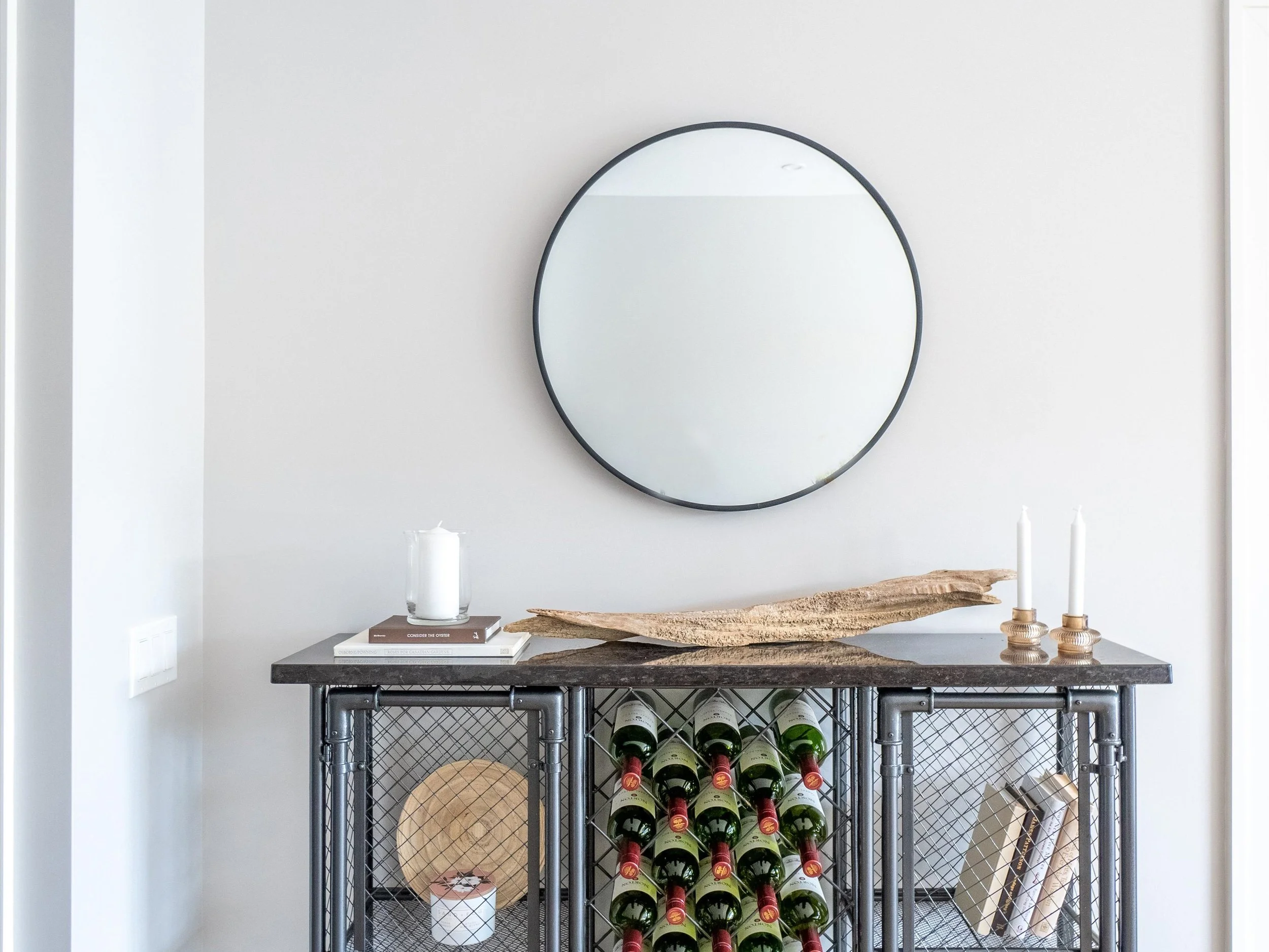 Modern wine storage unit with a black metal frame and a dark marble top, decorated with candles, a glass jar with a candle, a wooden art piece, books, and a round mirror on the plain white wall.  The Riverbend Condos, Huntsville, Muskoka.