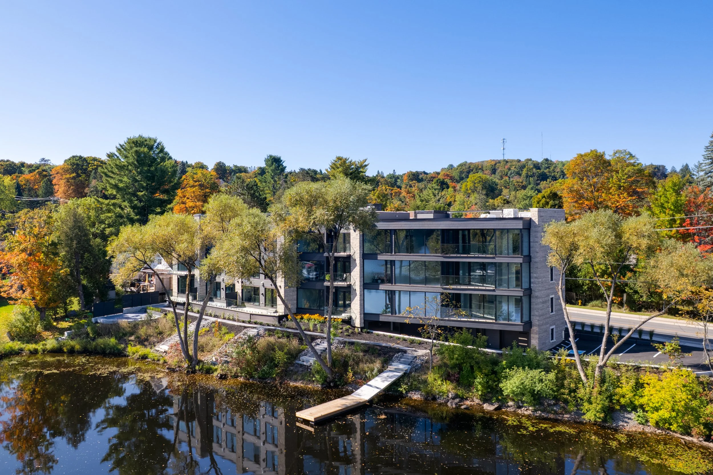 Modern multi-story apartment building beside a pond surrounded by colorful autumn trees with a hillside in the background.