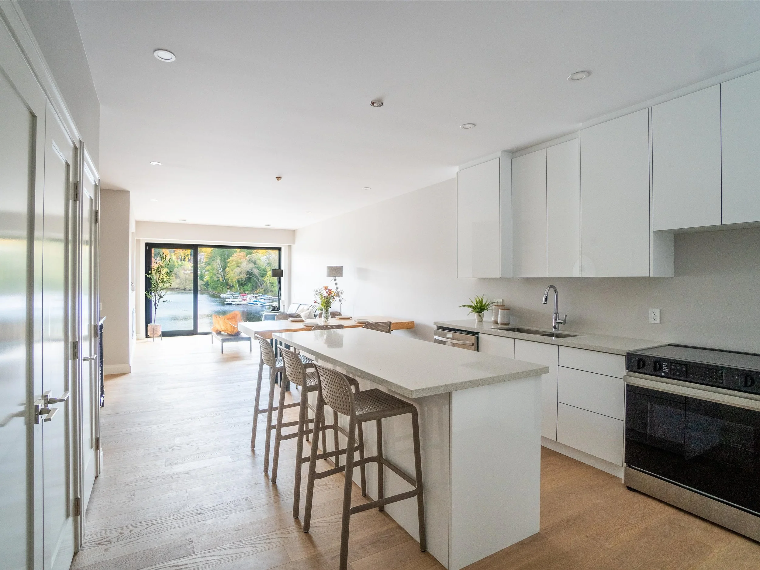 Modern open-concept apartment with kitchen, dining, and living area, large sliding glass door leading to balcony with water view, and minimalist decor.  The Riverbend Condos, Huntsville, Muskoka.