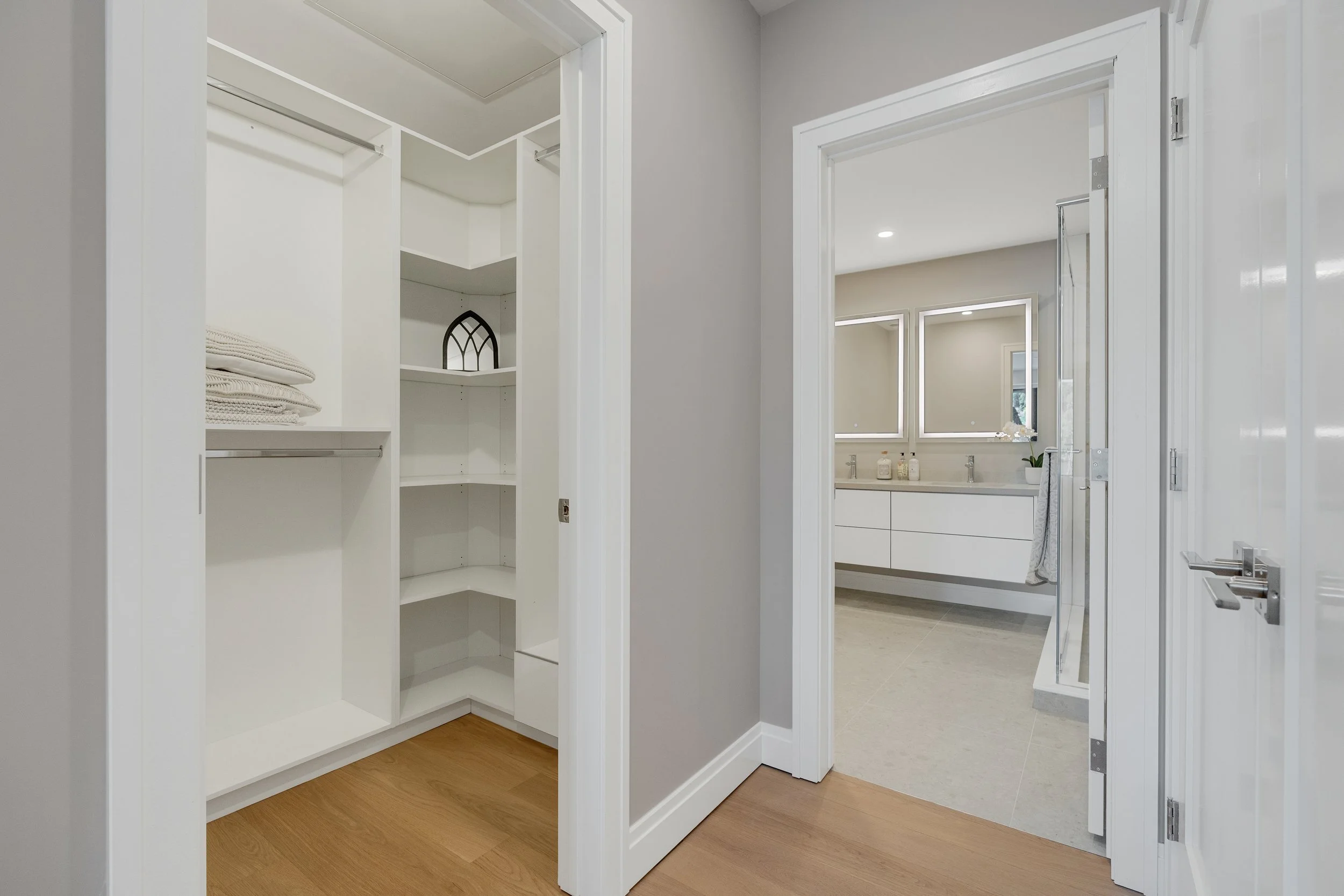 View of a walk-in closet with white shelving and a bathroom with a double vanity and two mirrors.  The Riverbend Condos, Huntsville, Muskoka.