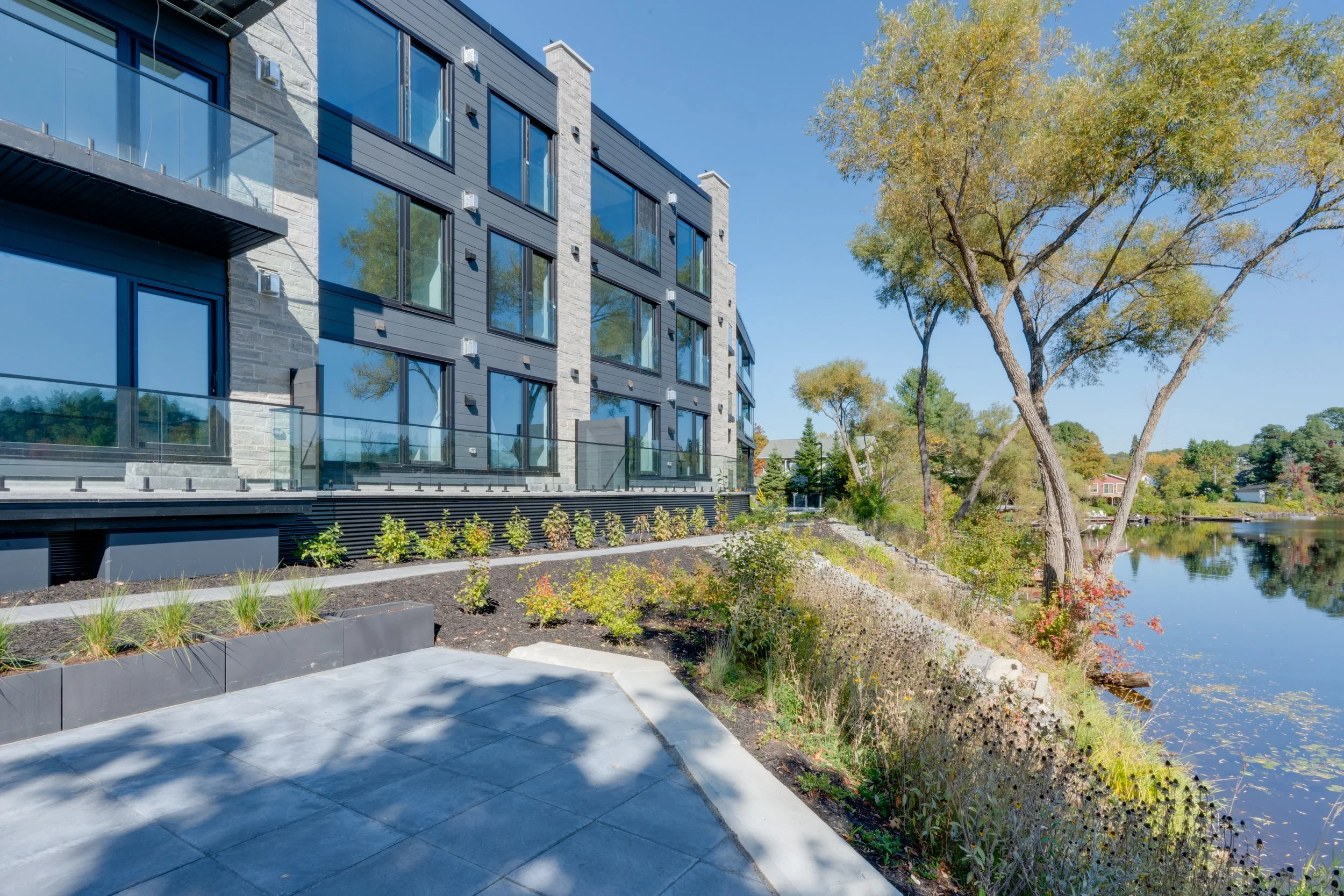 Modern multi-story apartment building with large windows and glass balconies beside a river, with trees and a clear blue sky in the background.