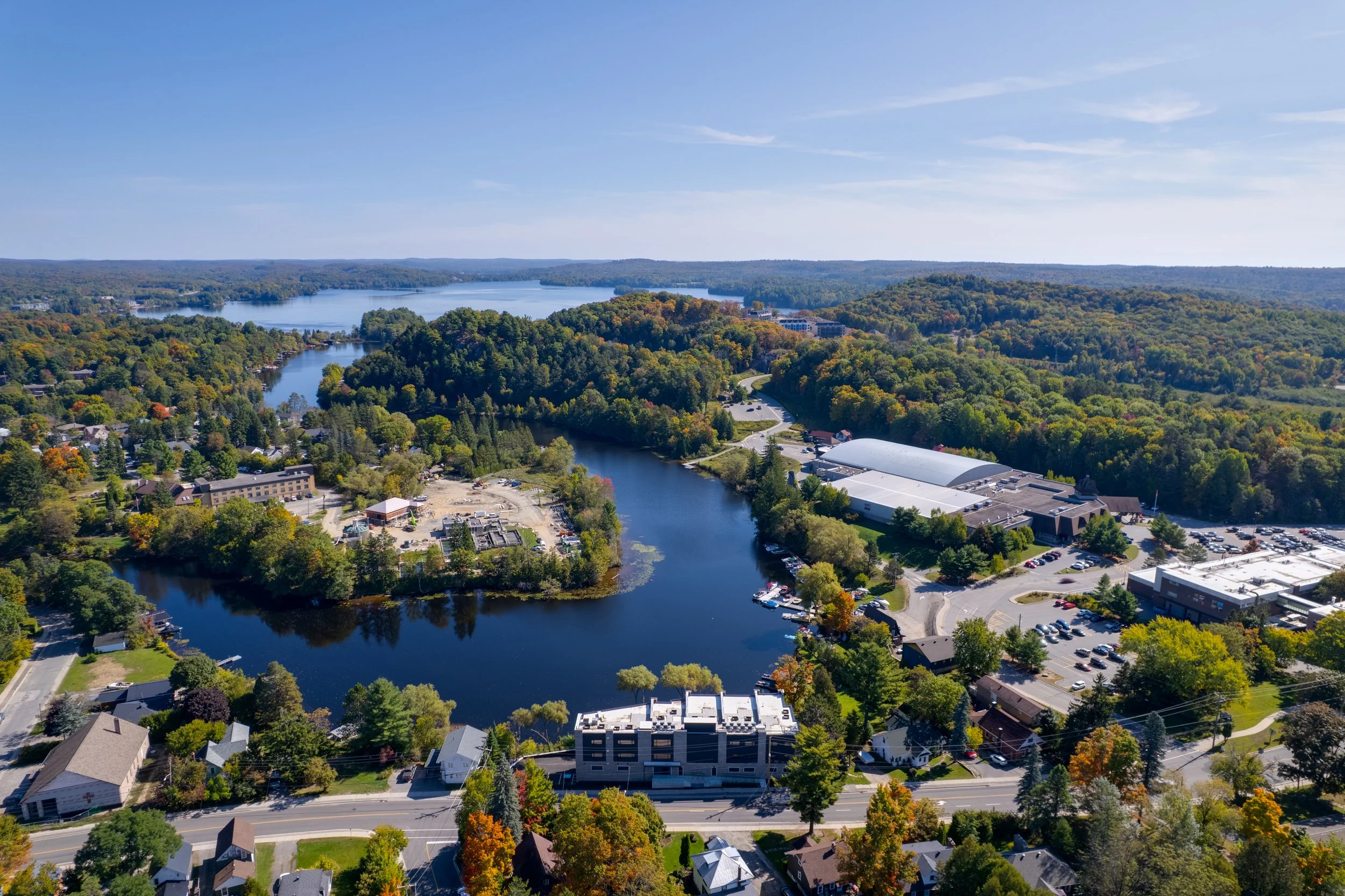 Aerial view of a lake surrounded by lush green trees, residential houses, commercial buildings, and parking areas under a clear blue sky.