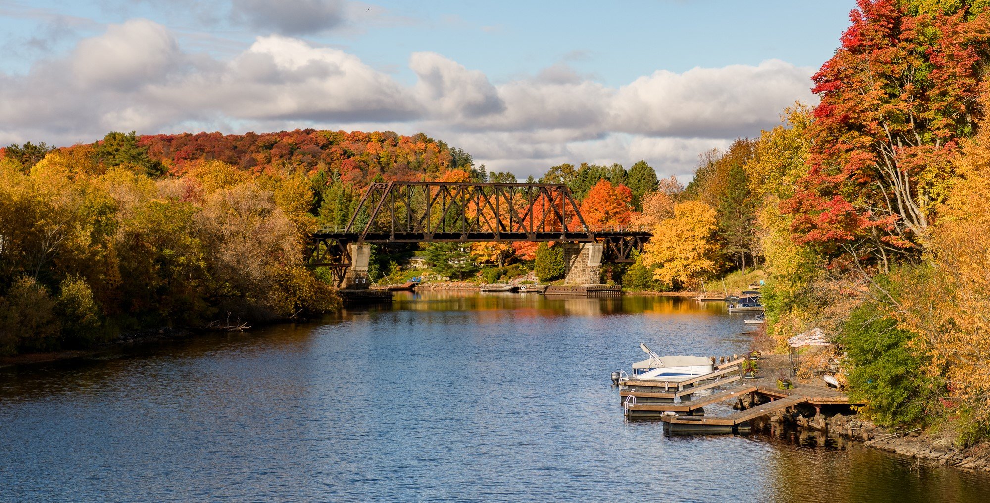 A scenic river view with a metal bridge crossing over the water, surrounded by colorful autumn trees with leaves in red, orange, and yellow. Several boats are docked along the shoreline.  The Riverbend Condos, Huntsville, Muskoka.