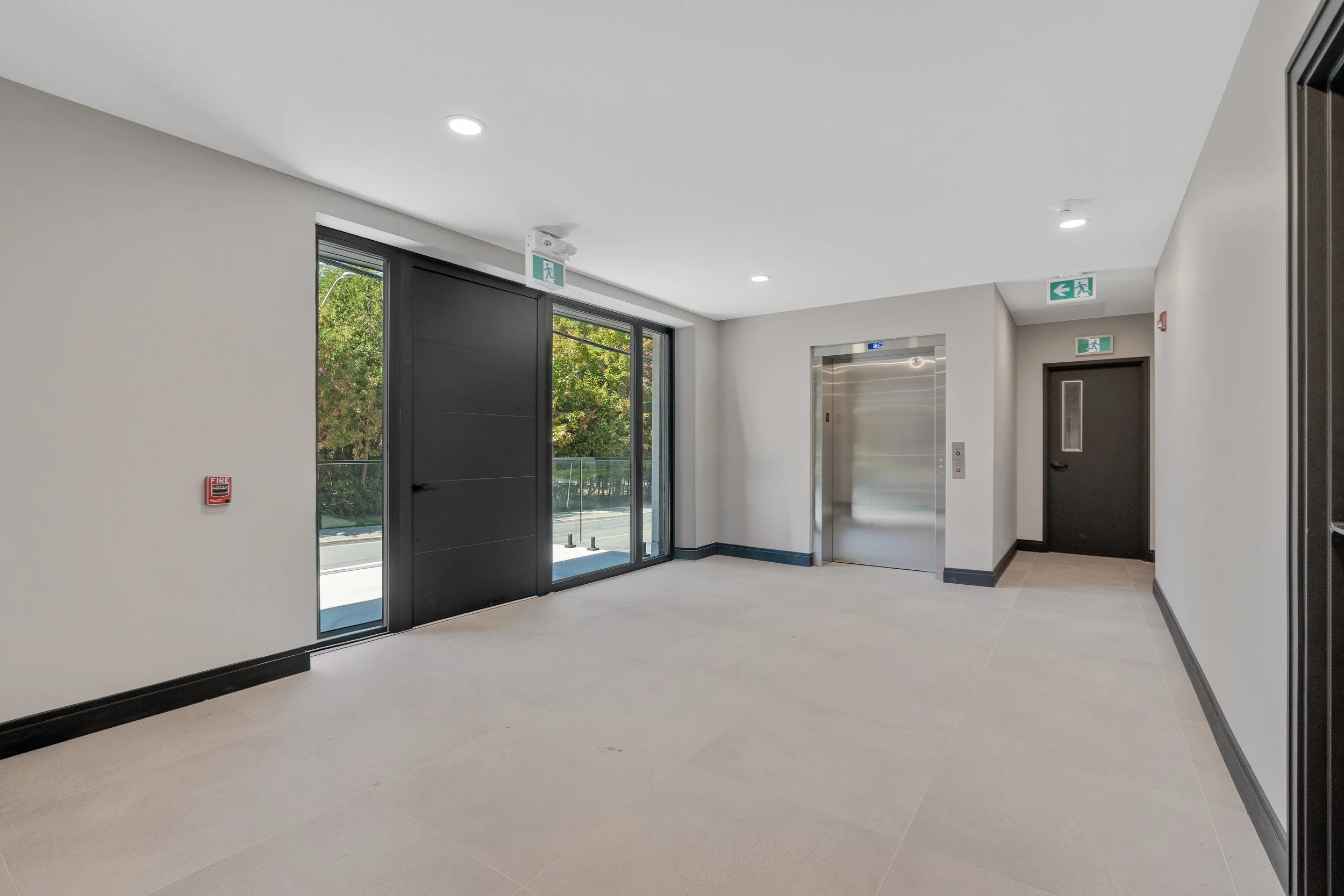 Interior of a modern building lobby with glass door entrance, elevator, emergency exit signs, and fire alarm, with a light-colored tiled floor and white walls.  The Riverbend Condos, Huntsville, Muskoka.