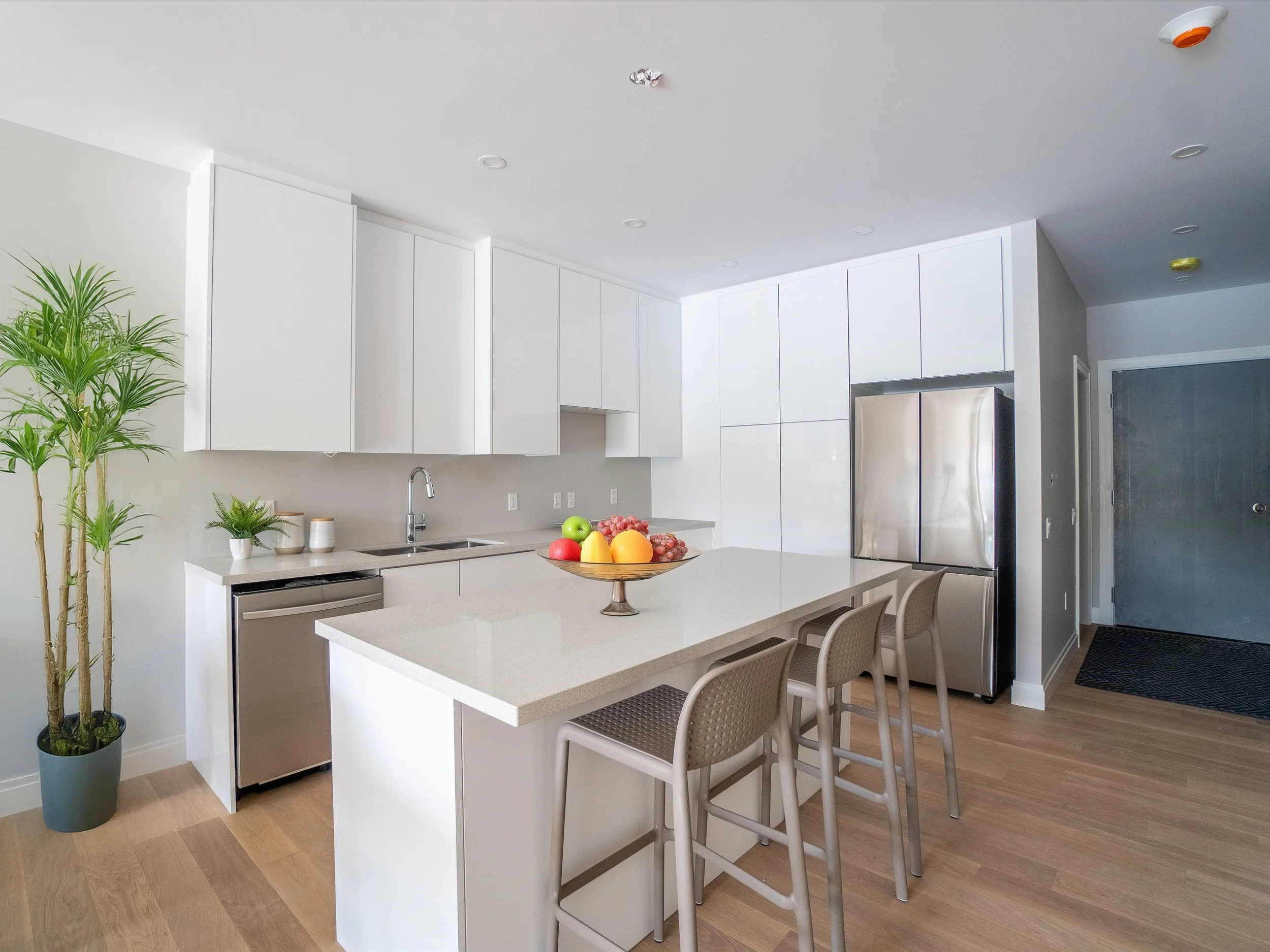 Modern kitchen with white cabinets, stainless steel refrigerator, wooden flooring, and a kitchen island with a bowl of assorted fruits.  The Riverbend Condos, Huntsville, Muskoka.