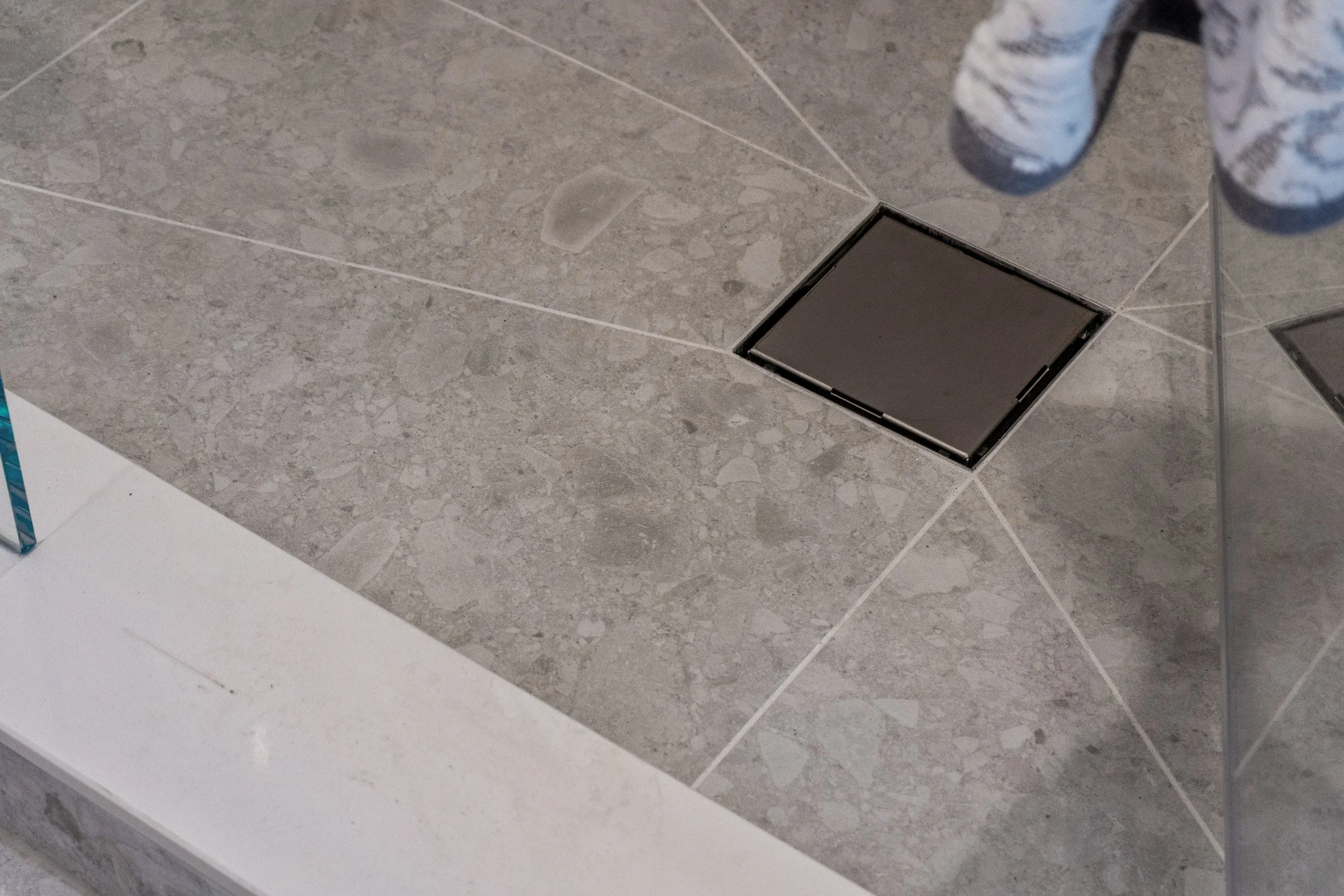 Gray tiled bathroom floor with a black floor drain and a person wearing white and gray striped slippers visible in the corner.  The Riverbend Condos, Huntsville, Muskoka.