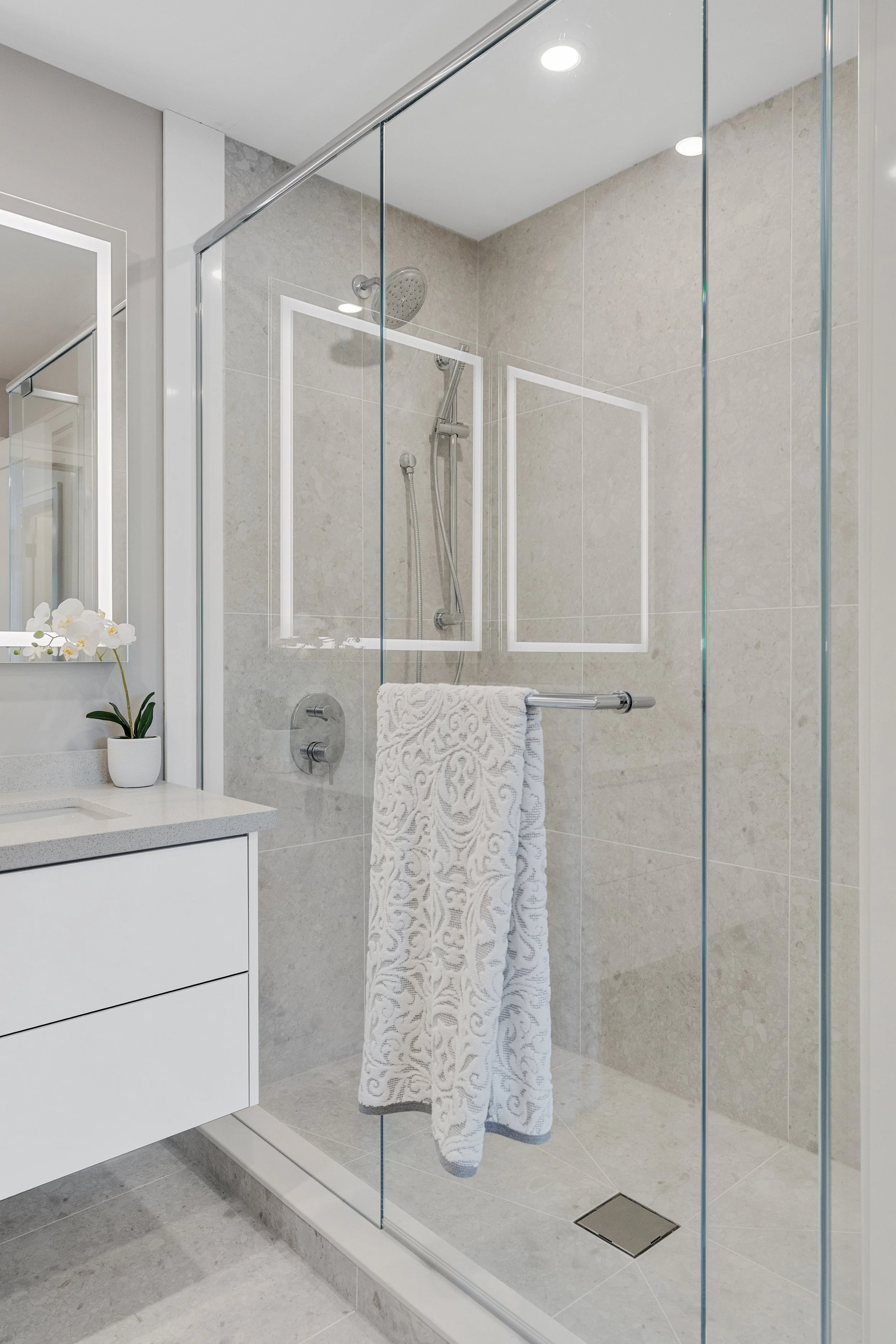 Modern bathroom with walk-in shower enclosed by glass. The shower has a rain showerhead and a hand-held shower. There is a towel hanging on a grab bar inside the shower.  The Riverbend Condos, Huntsville, Muskoka.