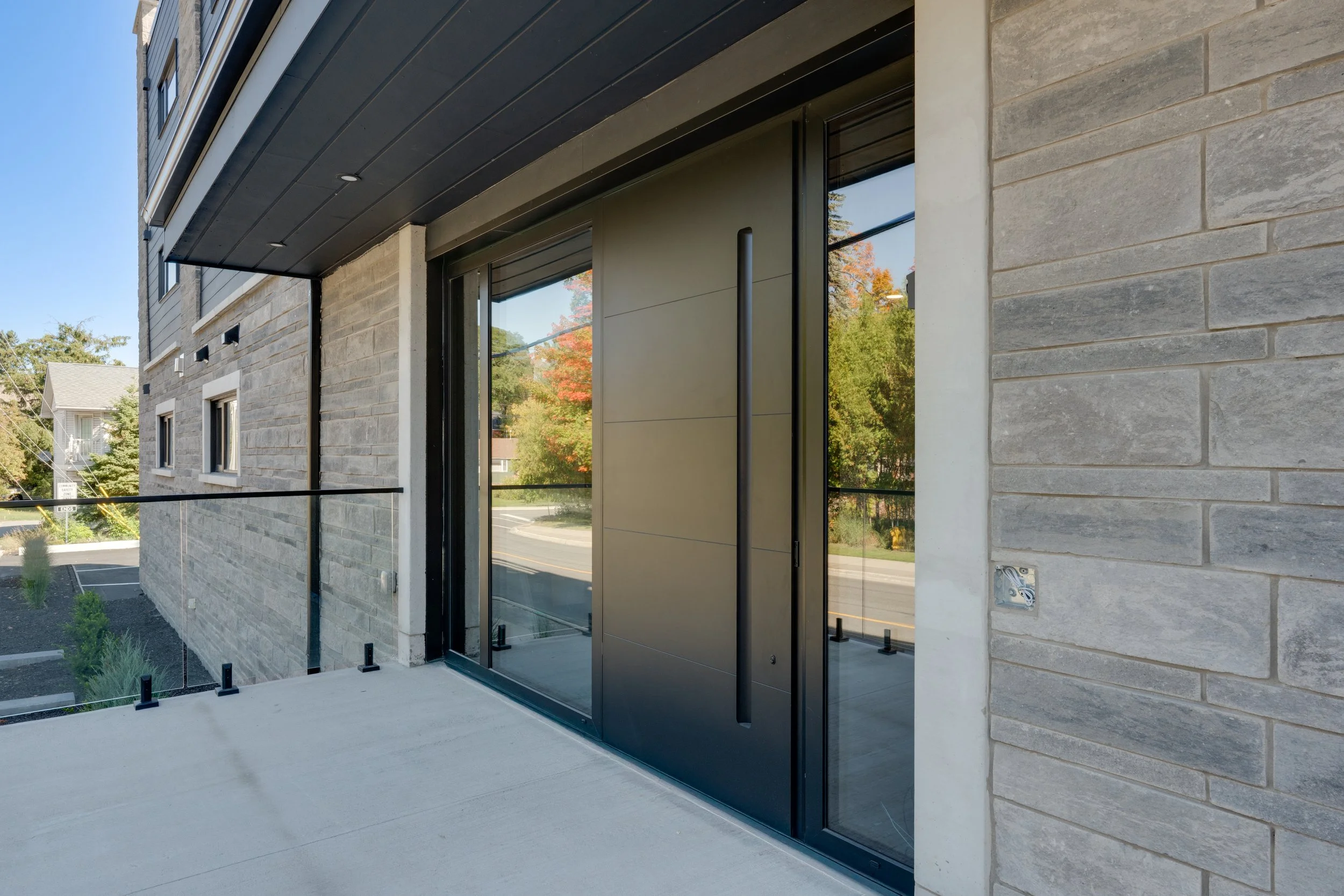 Modern building entrance with large glass sliding door, gray brick exterior, and a small balcony with black metal railing.  The Riverbend Condos, Huntsville, Muskoka.