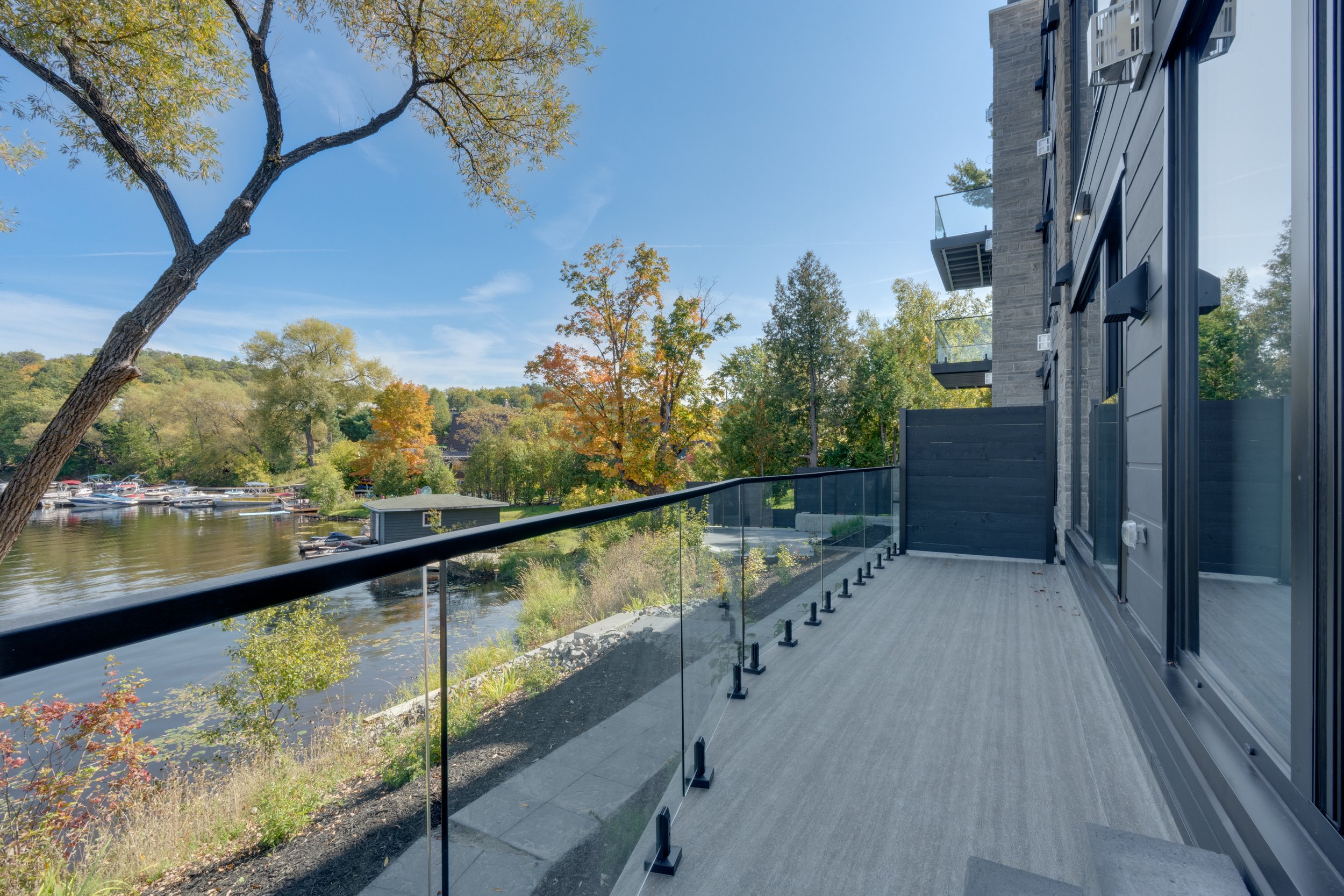 A balcony overlooking a river with boats, surrounded by trees with fall colors, next to a modern building with large windows.