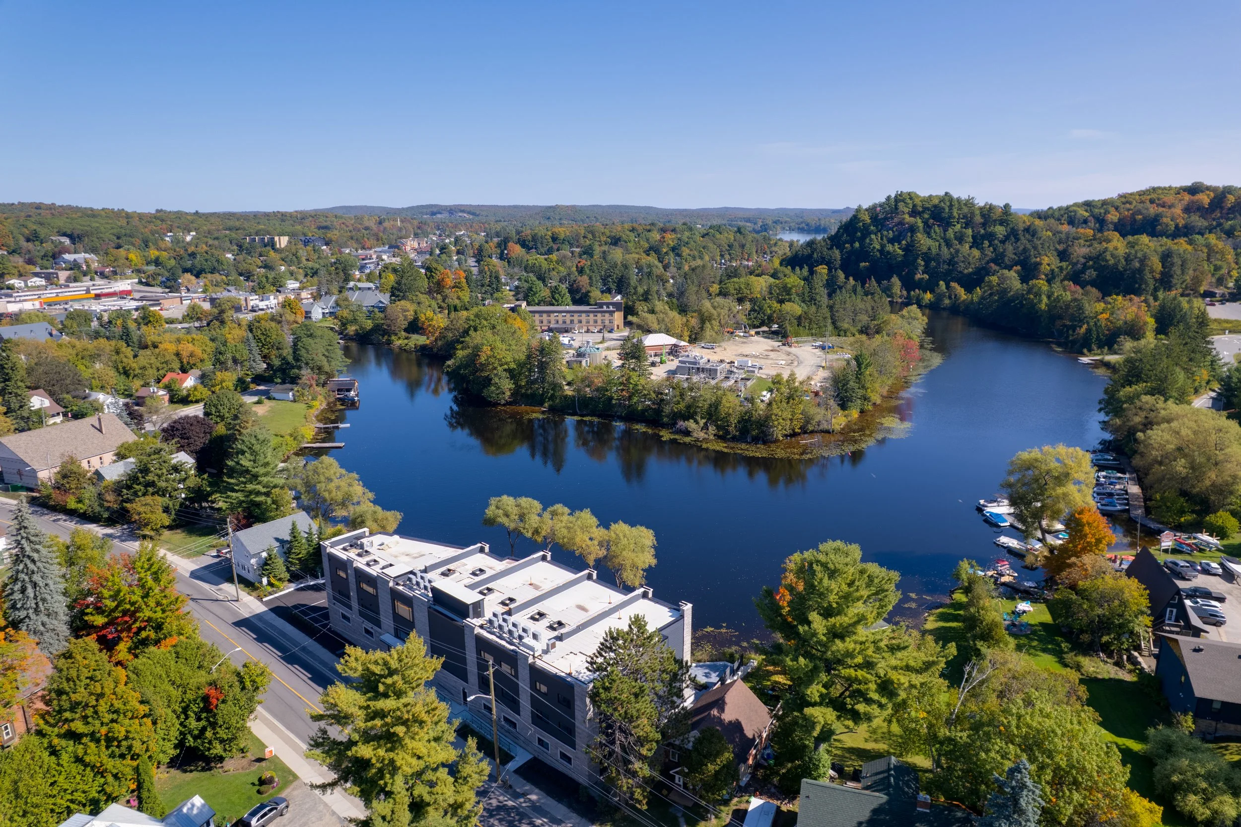 Aerial view of a river surrounded by trees and buildings in a small town on a clear day.  The Riverbend Condos, Huntsville, Muskoka.