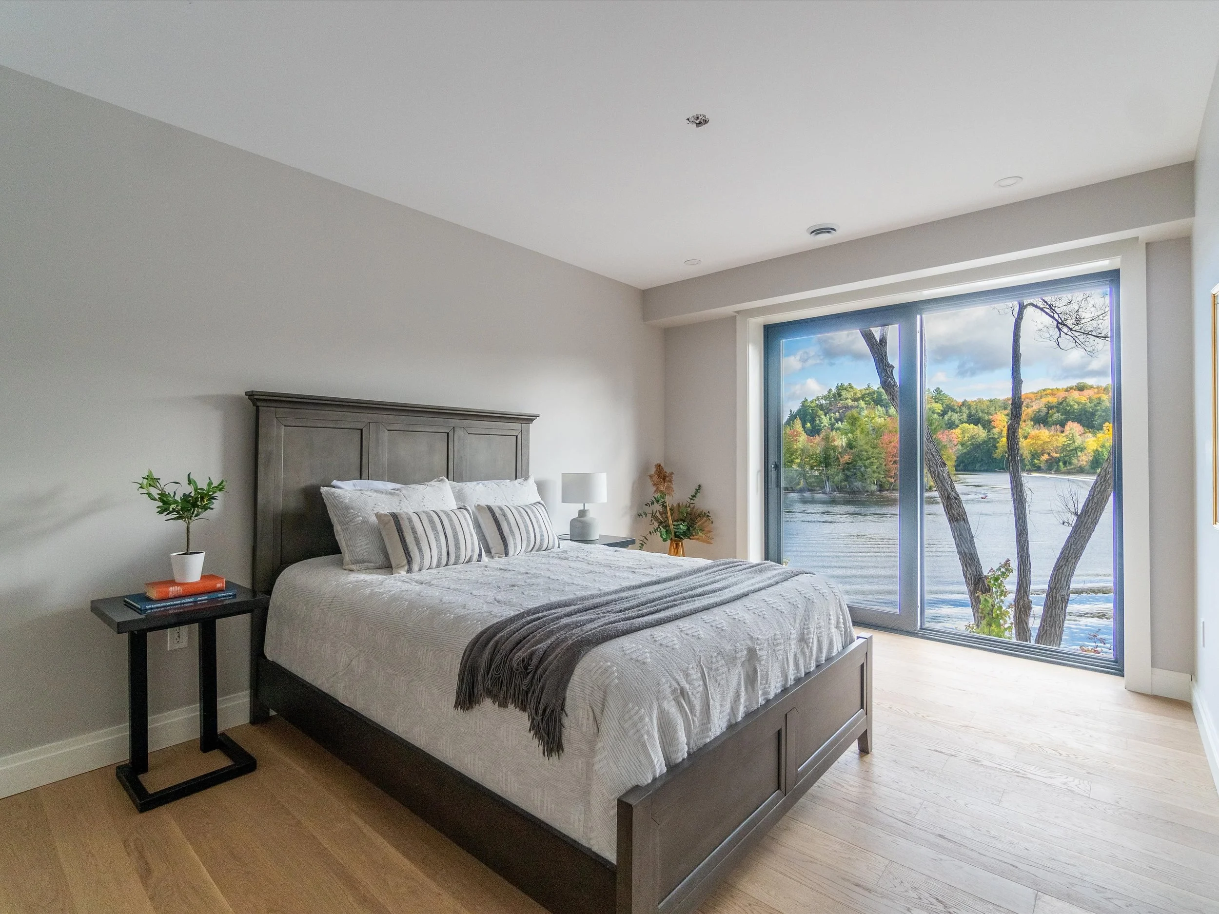 Bedroom with large window showing a river and trees with fall foliage, bed with gray headboard, white bedding, and gray throw blanket, nightstand with plant, lamp, and books.  The Riverbend Condos, Huntsville, Muskoka.