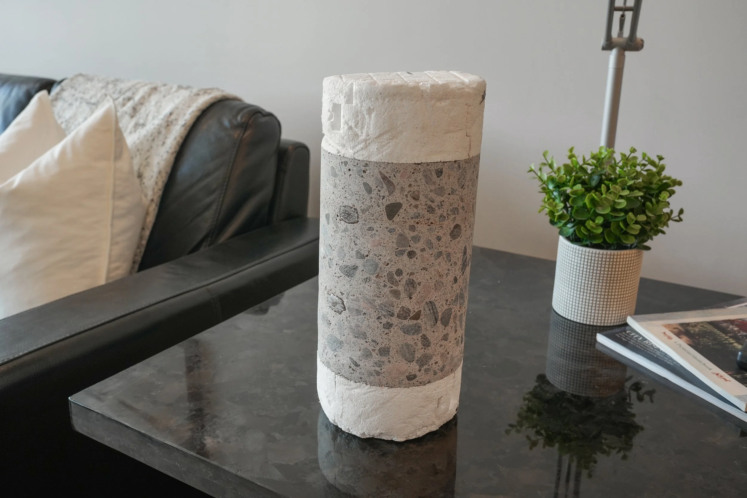Decorative cylindrical tabletop sculpture made of white stone and concrete on a black marble surface, with a potted green plant and magazines in the background.  The Riverbend Condos, Huntsville, Muskoka.