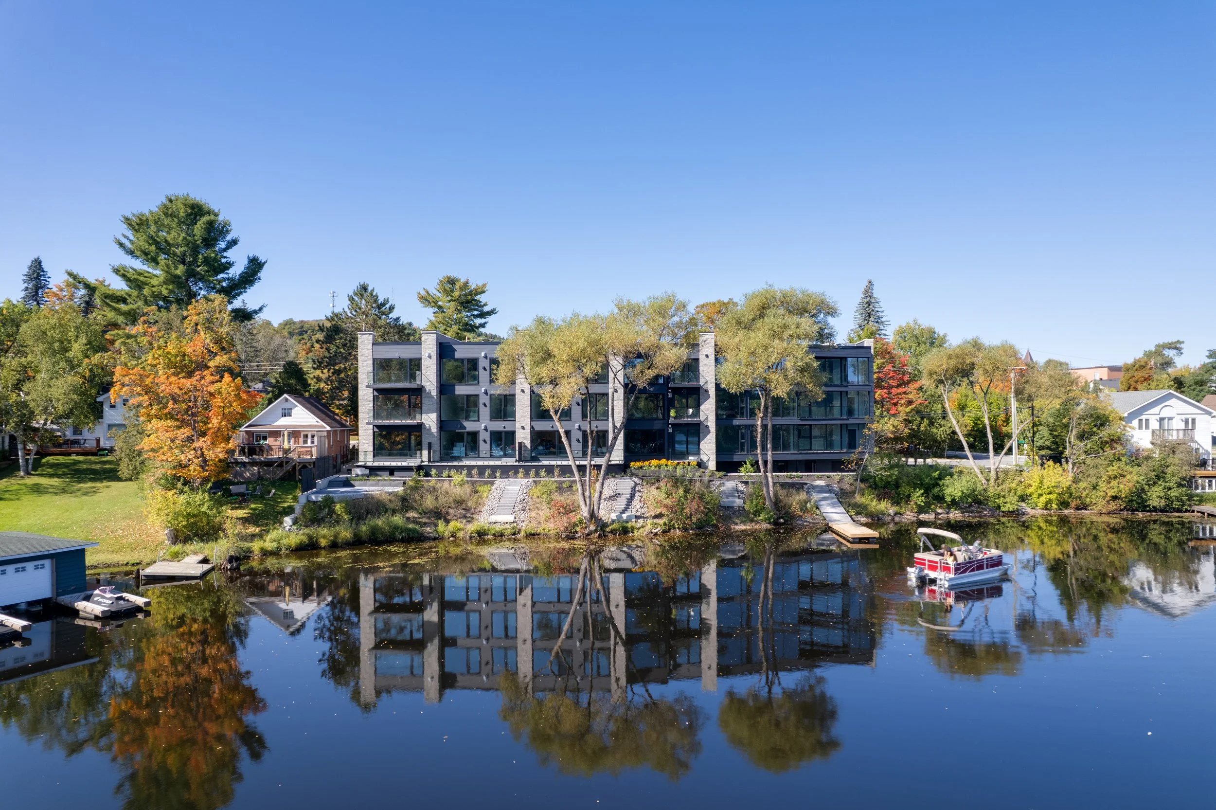 Modern multi-story apartment building by a river, with trees and houses around, a boat in the water, and a clear blue sky.