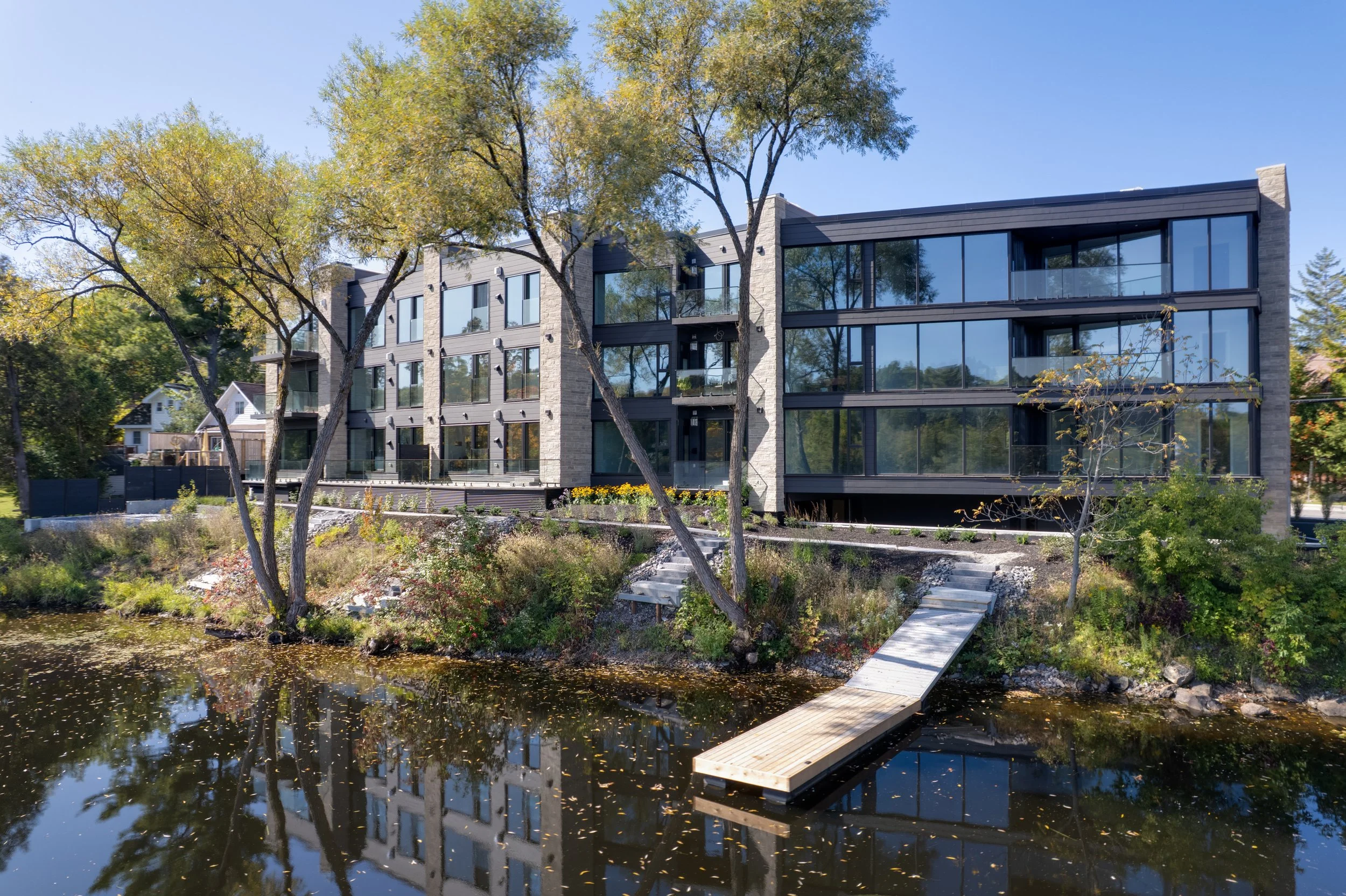 Modern multi-story apartment building with large glass windows, situated along a pond with a wooden dock, trees, and landscaping. The Riverbend Condos, Huntsville, Muskoka.